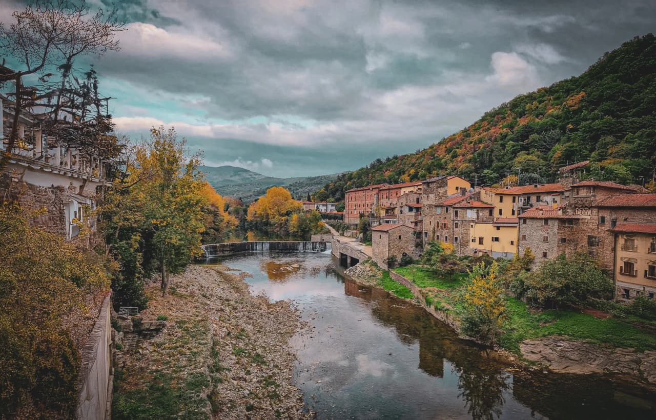 A picturesque Tuscan village bordered by a tranquil river, surrounded by green hills. Stone buildings with colourful facades stand alongside the river, with red-tiled roofs.