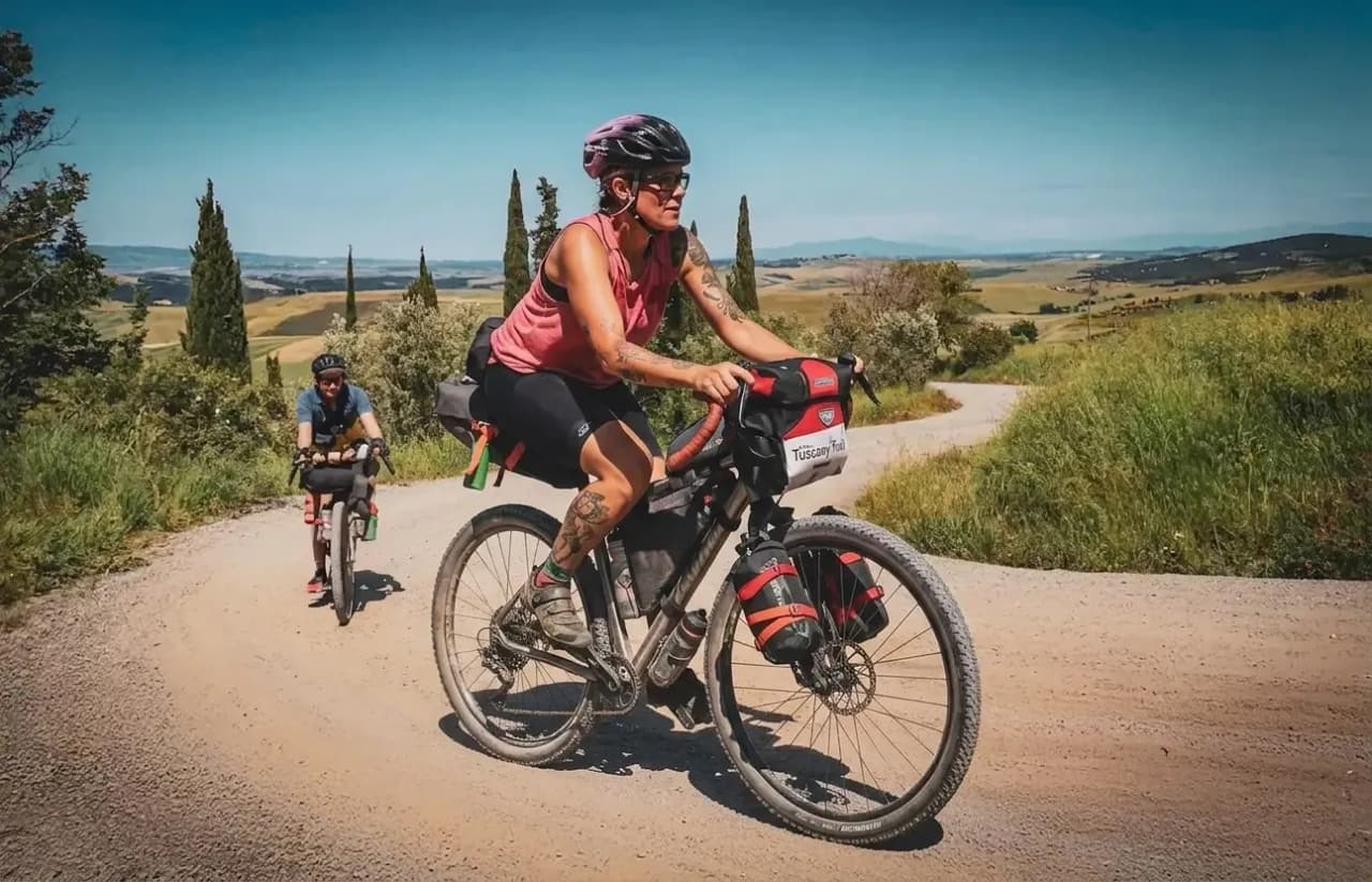 Un couple de cyclistes évolue sur un chemin de gravier sinueux, entouré d'une nature verdoyante et de collines ondulantes. L'un des cyclistes, en maillot rose et casque.