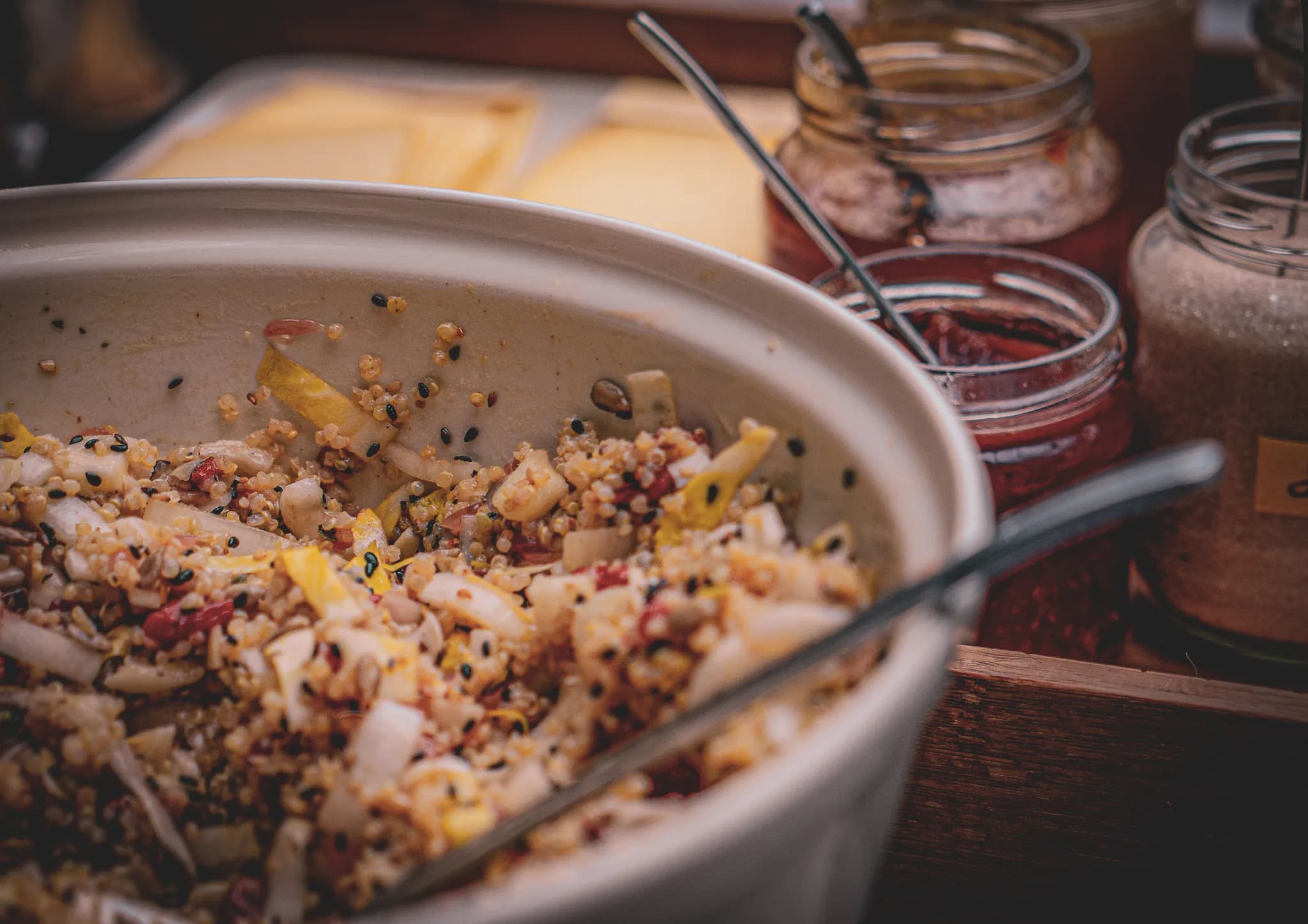 A dish of quinoa is filled with a variety of vegetables, such as pieces of yellow pepper and onion, as well as seeds. In the background, a number of glass jars contain different condiments and ingredients, such as