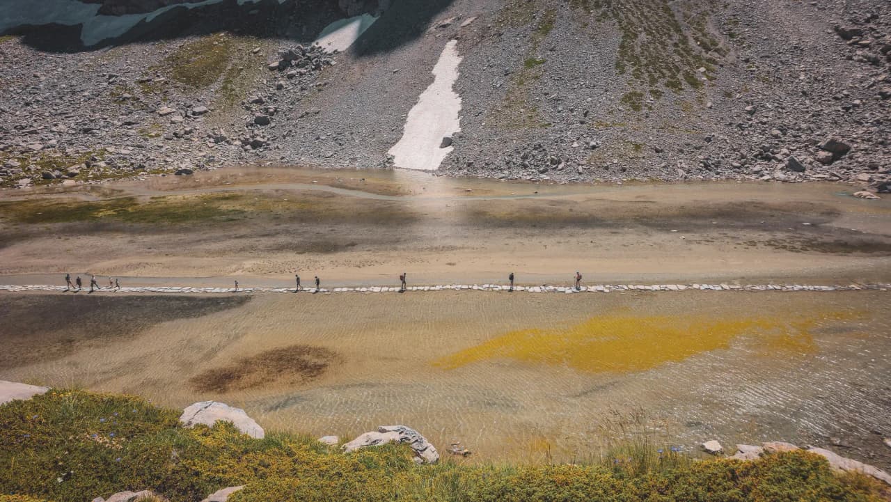 A group of hikers cross a high-altitude lake over a stone footbridge, surrounded by Alpine scenery.
