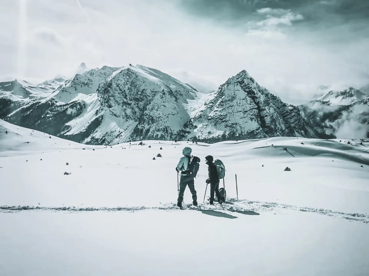Two hikers on snowshoes on a vast expanse of immaculate snow, facing majestic peaks.