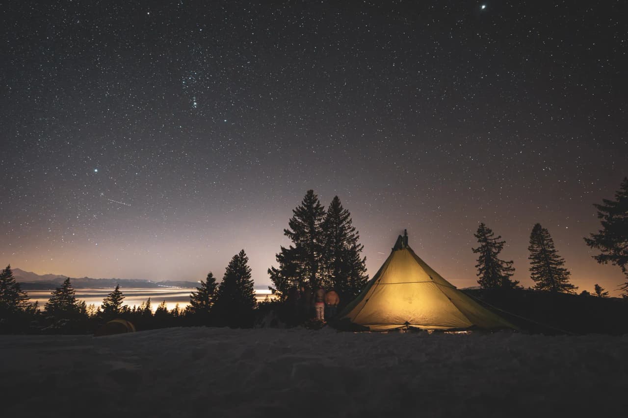Sous un ciel étoilé, une tente lumineuse se dresse dans la neige, entourée de majestueux sapins.