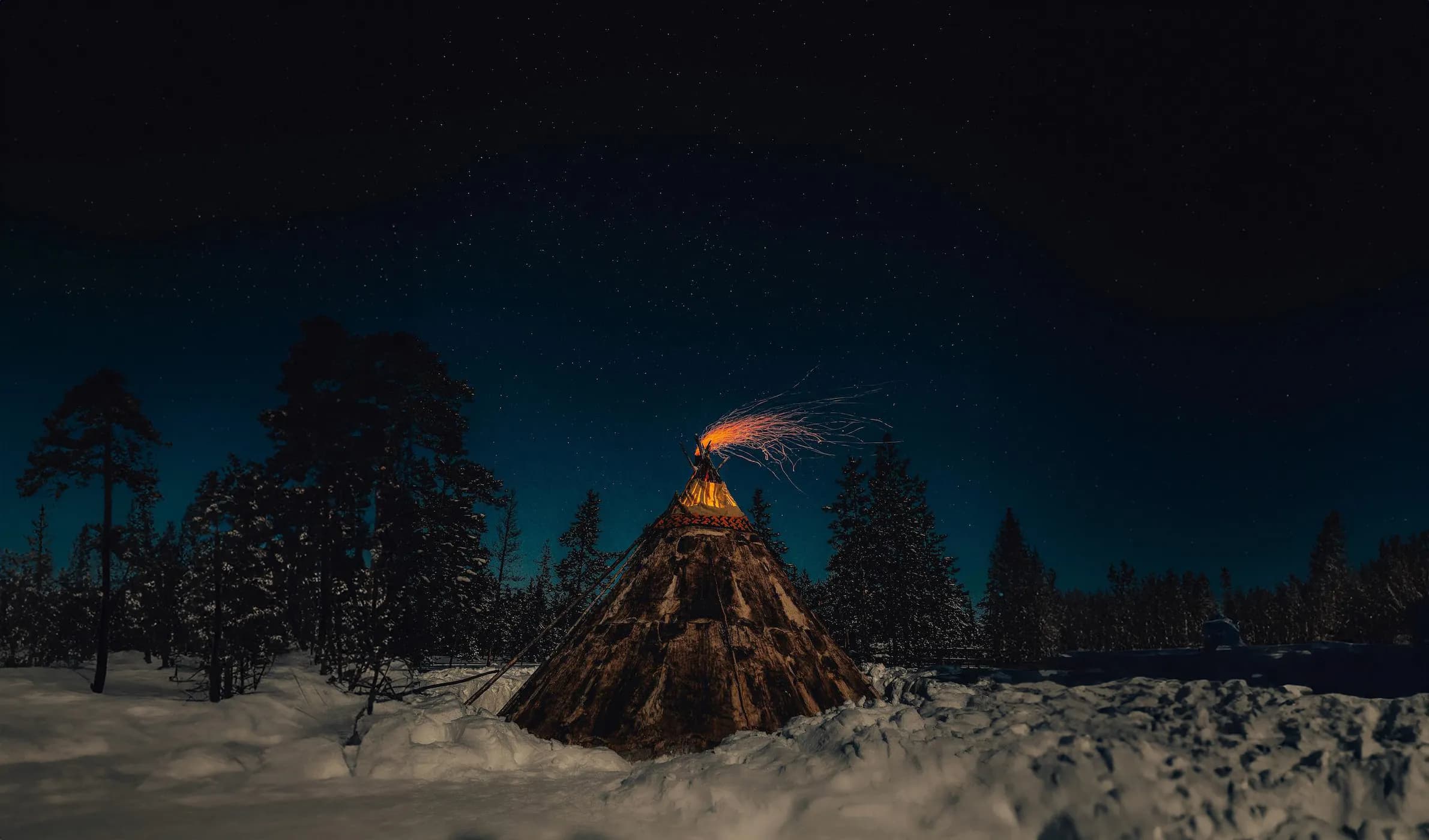 A wooden tipi, lit by a sparkling fire, stands in the middle of a snowy landscape. The starry night adds a peaceful, mystical atmosphere, while the fir trees
