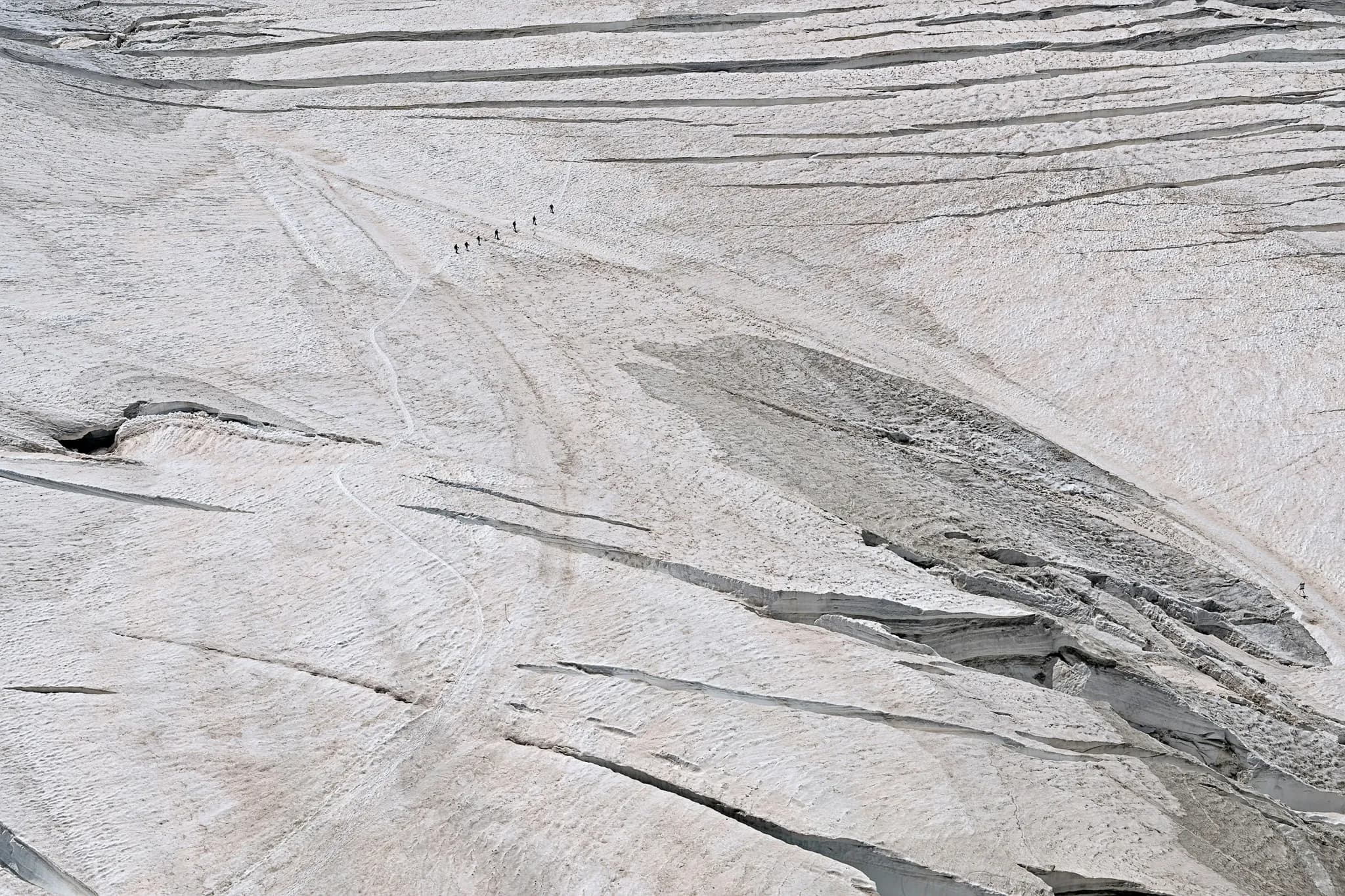 A vast and impressive glacier covers the landscape, with strata and fissures testifying to its history. In the distance, a group of people walk along a winding path.