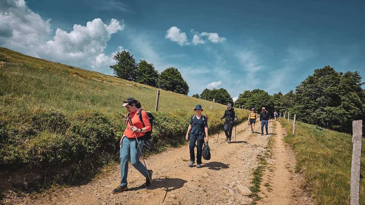 Two hikers are walking across a vast green meadow with hills under a cloudy sky.