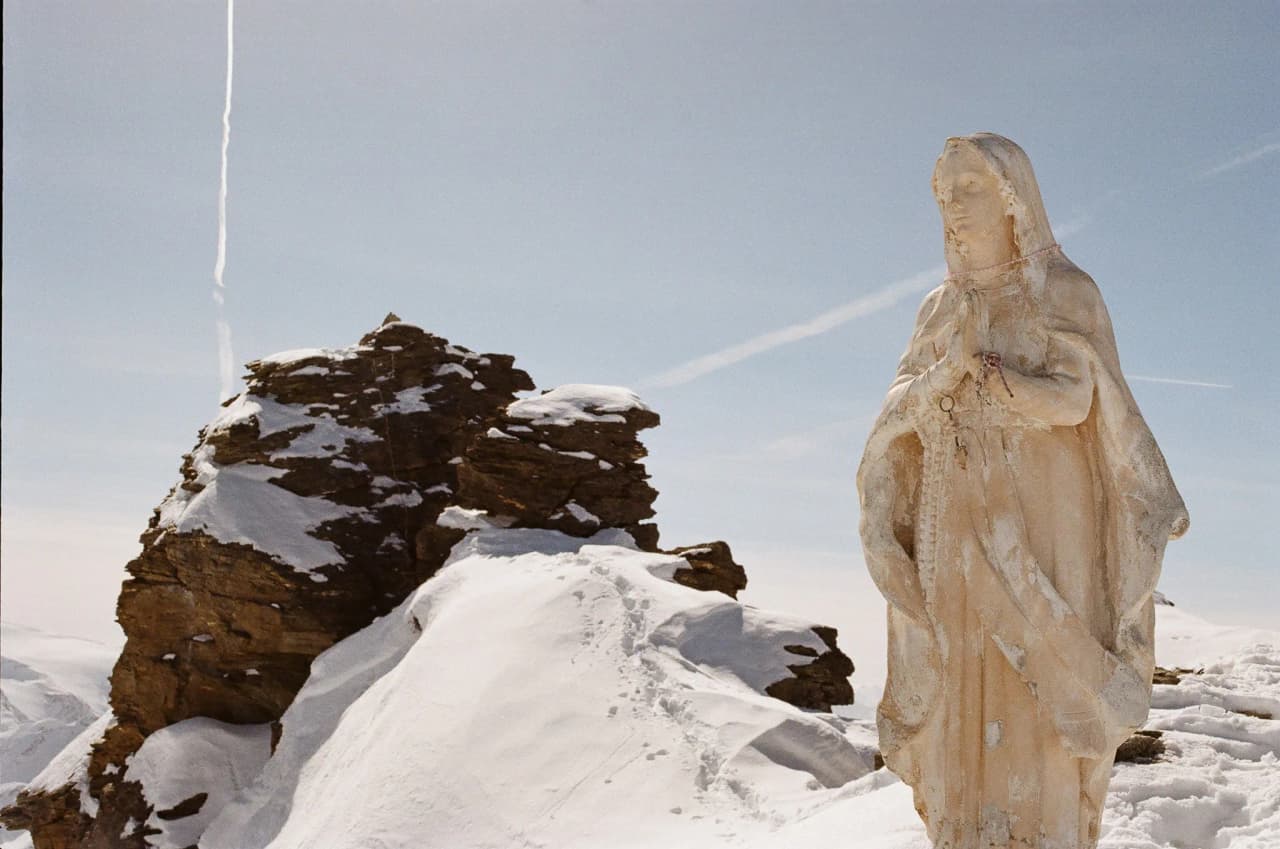 Une statue majestueuse se dresse sur un sommet alpin enneigé, avec des paysages à couper le souffle.