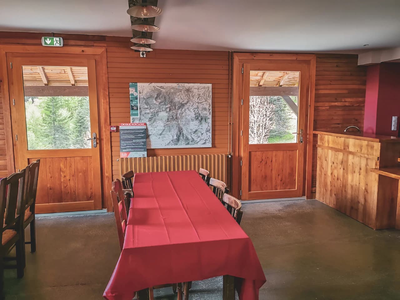 Intérieur chaleureux d'un refuge en bois, table rouge, vue sur la nature environnante.