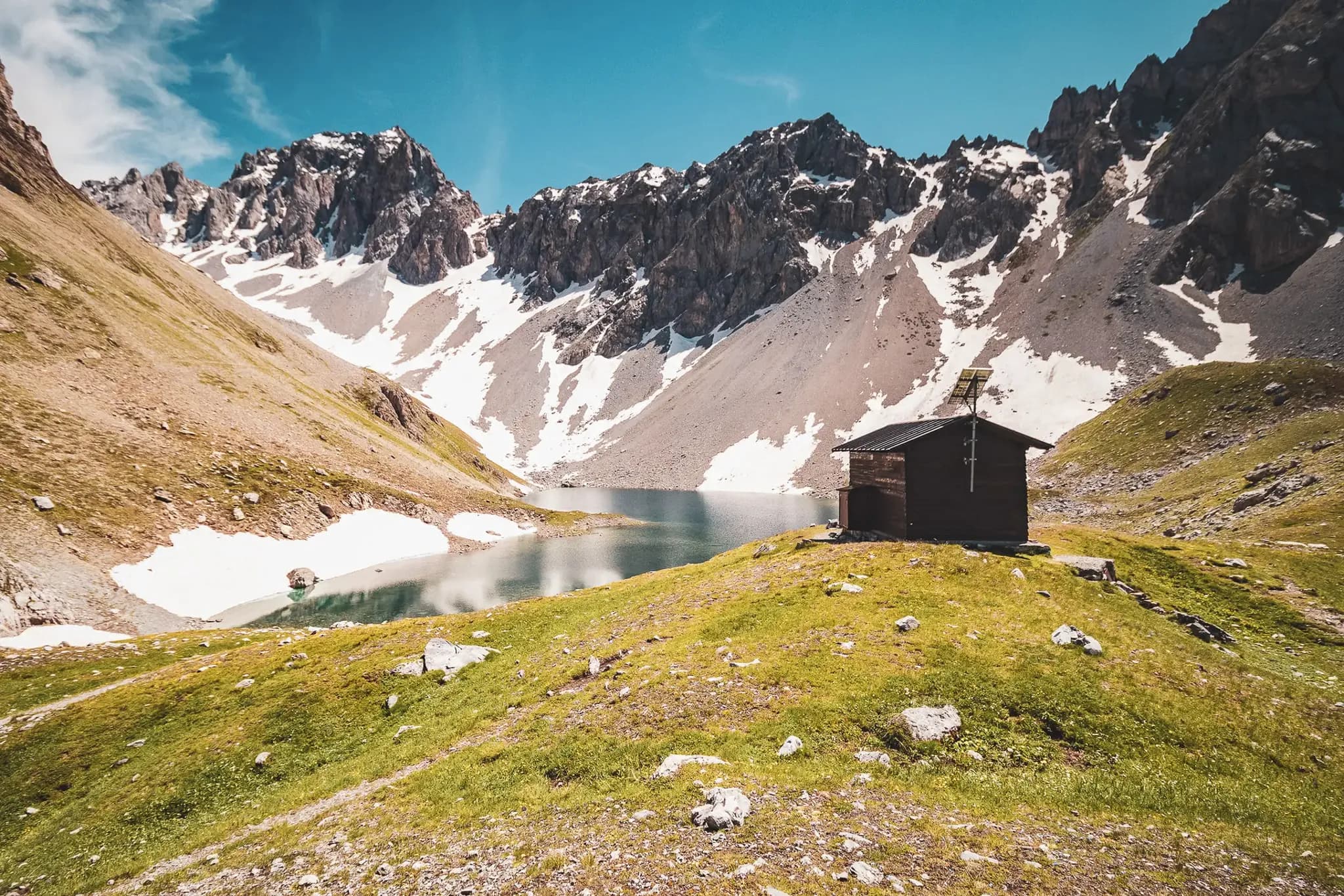 A wooden chalet stands on a green hillside beside a tranquil lake. The landscape is surrounded by majestic snow-capped mountains, under a clear blue sky. The grass is lush