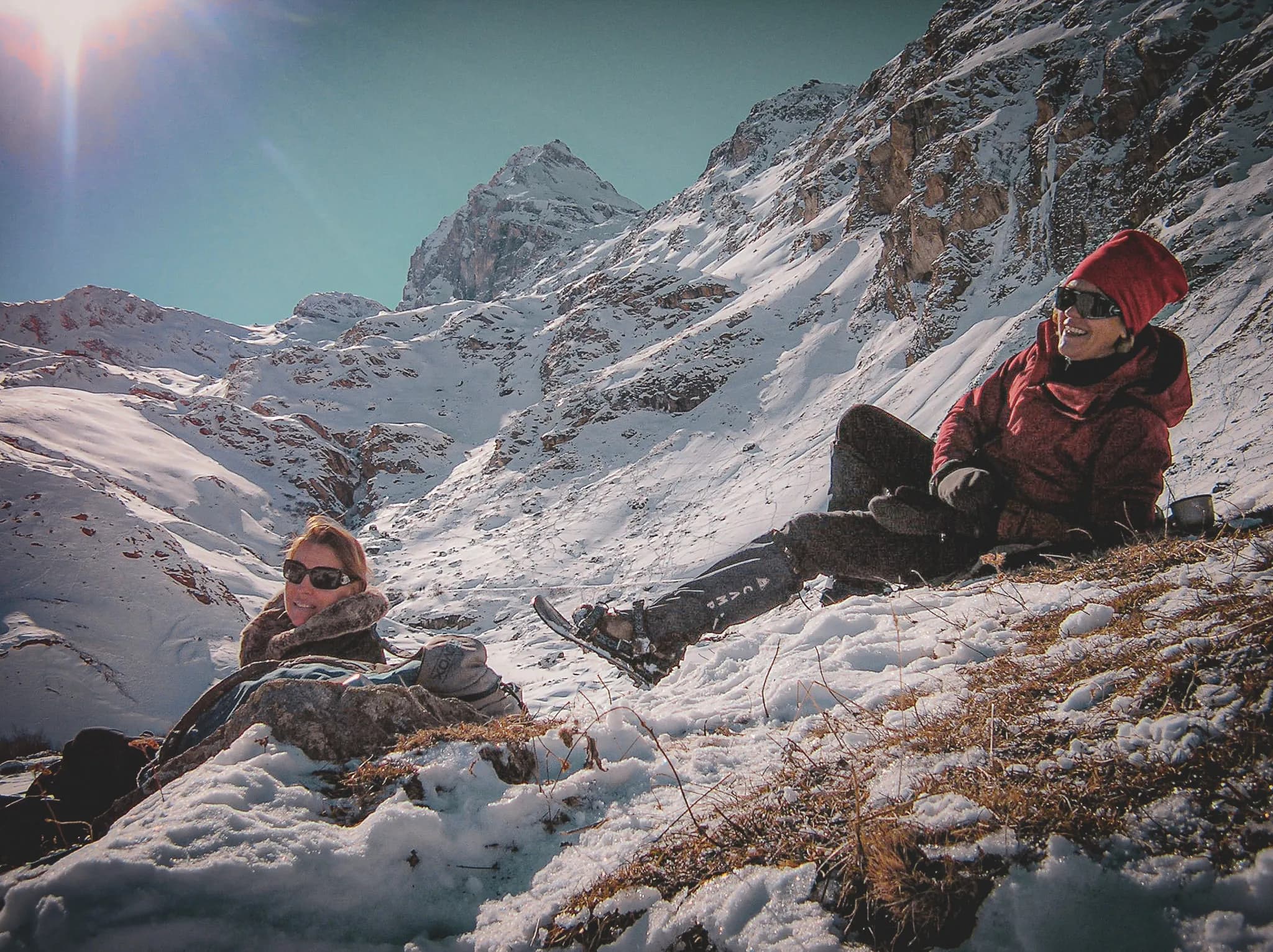 Two people are resting in a snowy alpine landscape, surrounded by majestic mountains. One of them is lying on the snow, smiling, while the other is sitting next to her, dressed in a