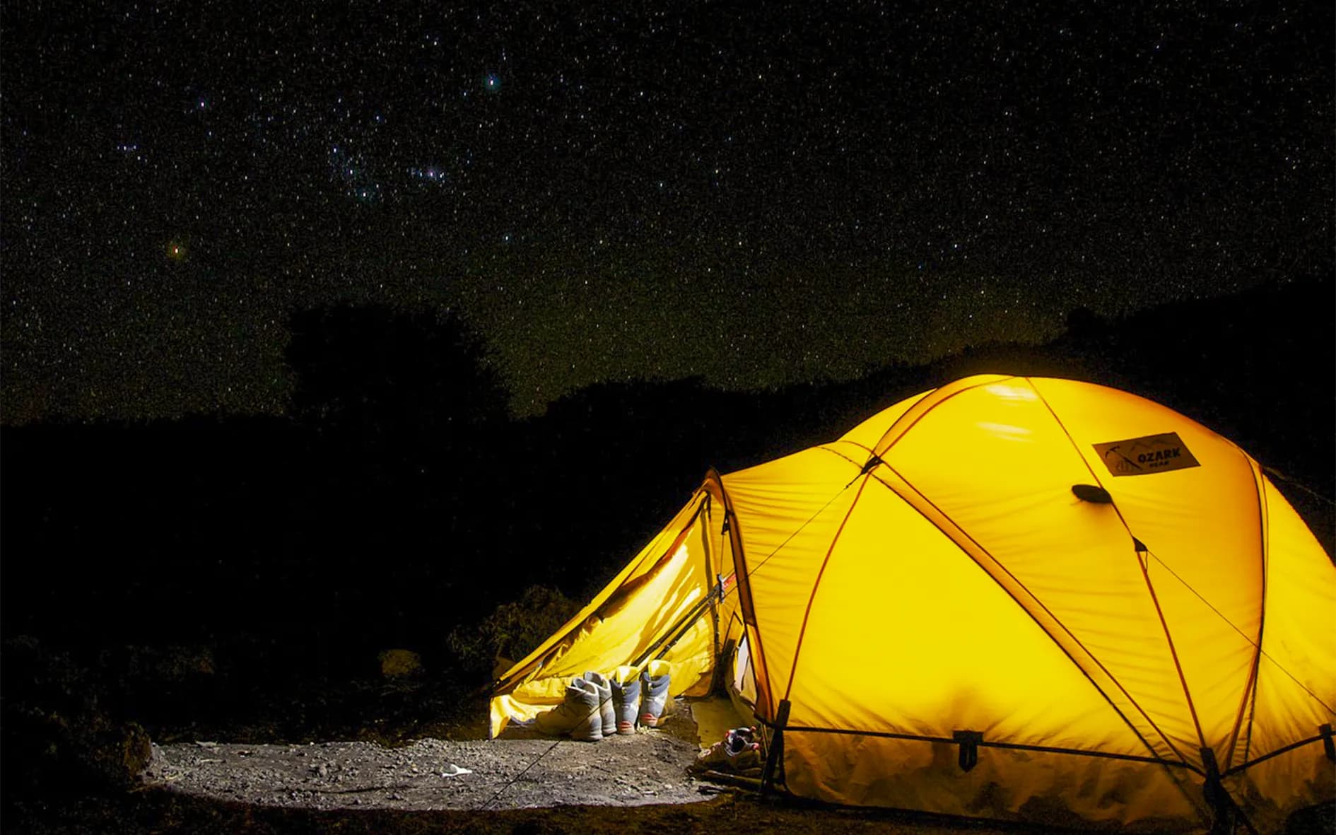 A bright yellow tent is set up under a starry sky, creating a soothing and adventurous atmosphere. Shoes can be seen in front of the entrance to the tent, on an earthen floor, adding to the atmosphere.