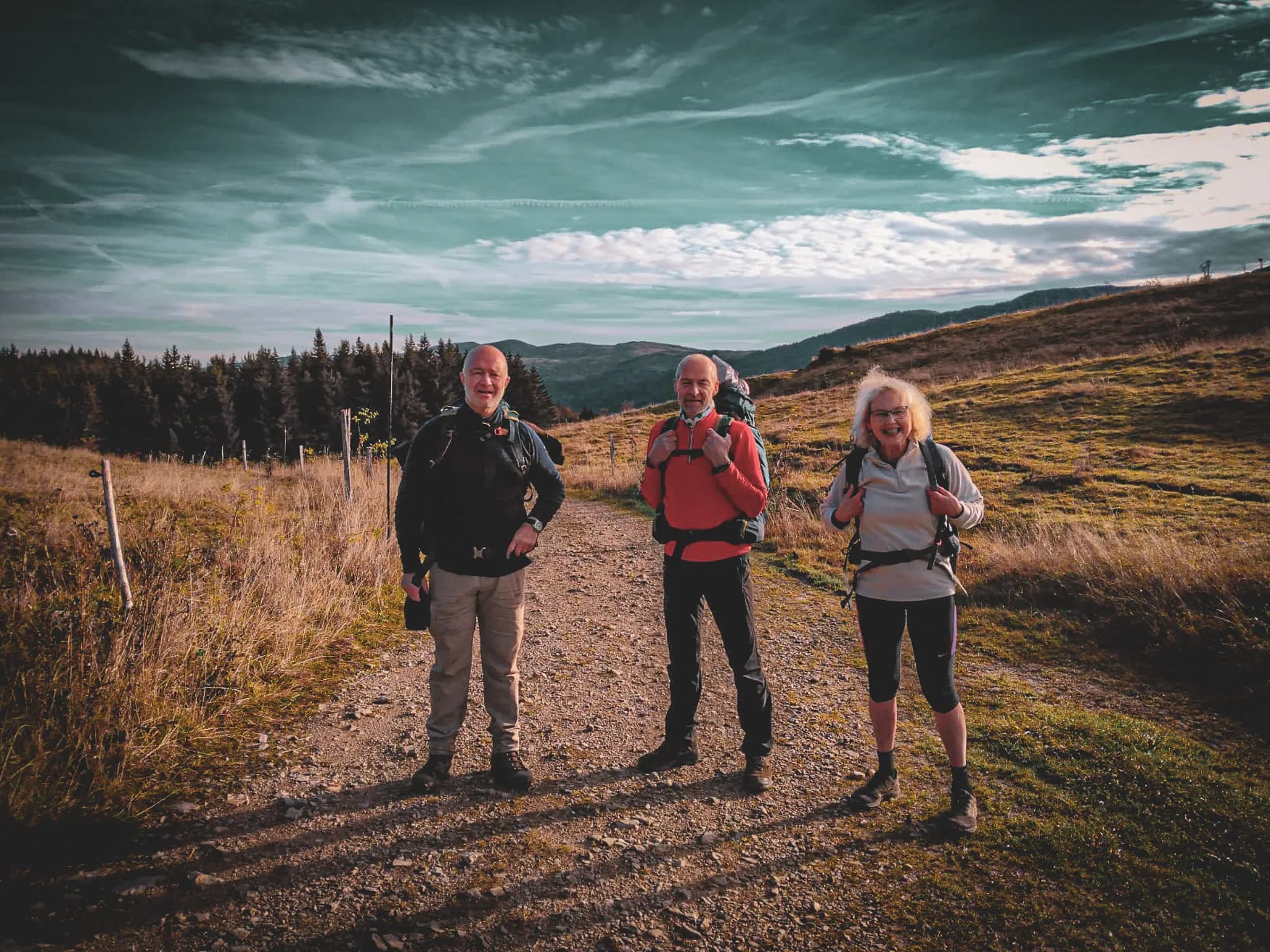 Three hikers are on a dirt track surrounded by natural scenery. Two men and a woman, all carrying rucksacks, seem to be enjoying a day in the great outdoors. The man on the left has a grey beard and