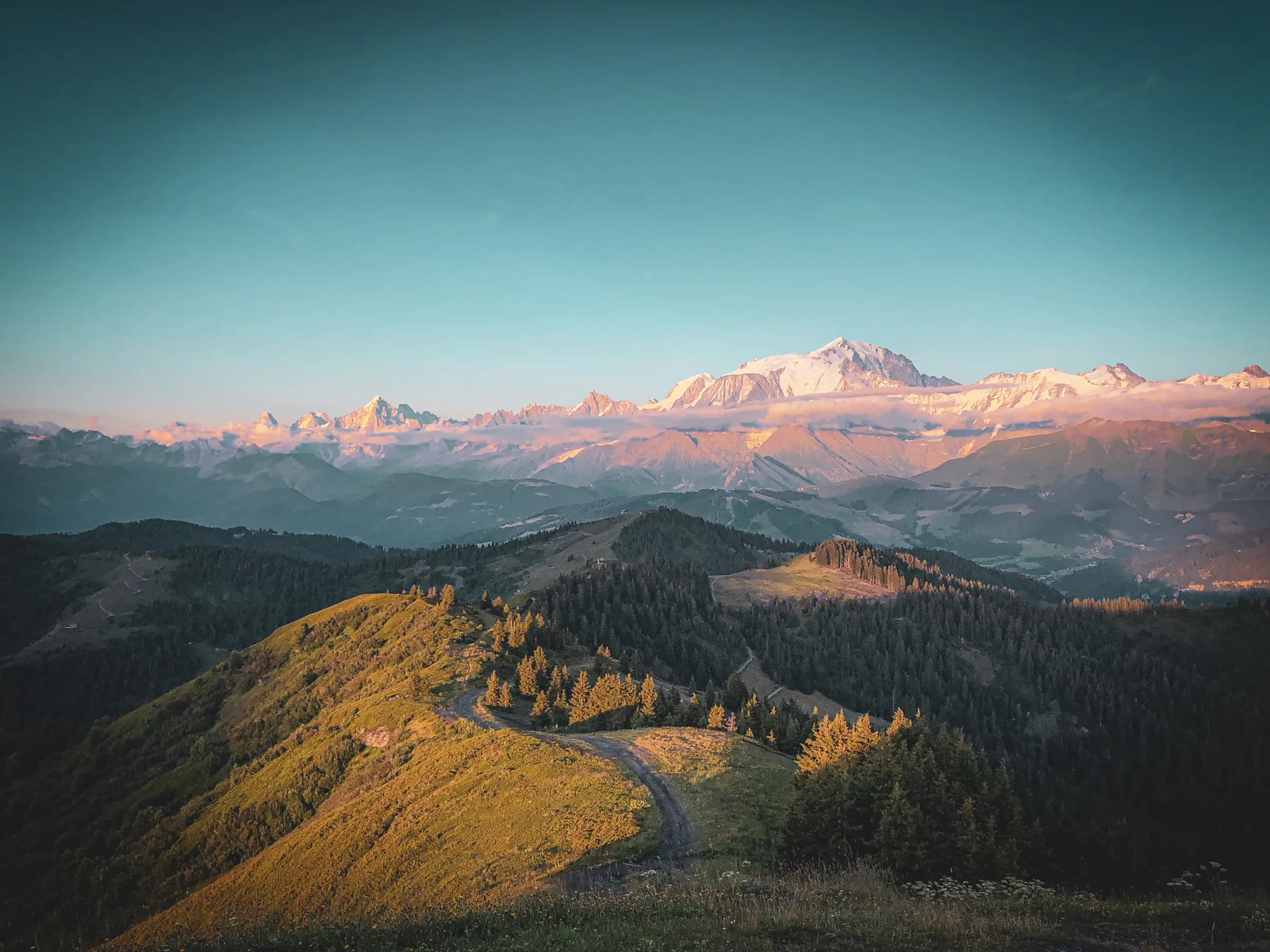 Spectacular mountain scenery stretches as far as the eye can see, with majestic snow-capped mountains illuminated by the soft light of dusk. In the foreground, lush green hills are dotted across the landscape.