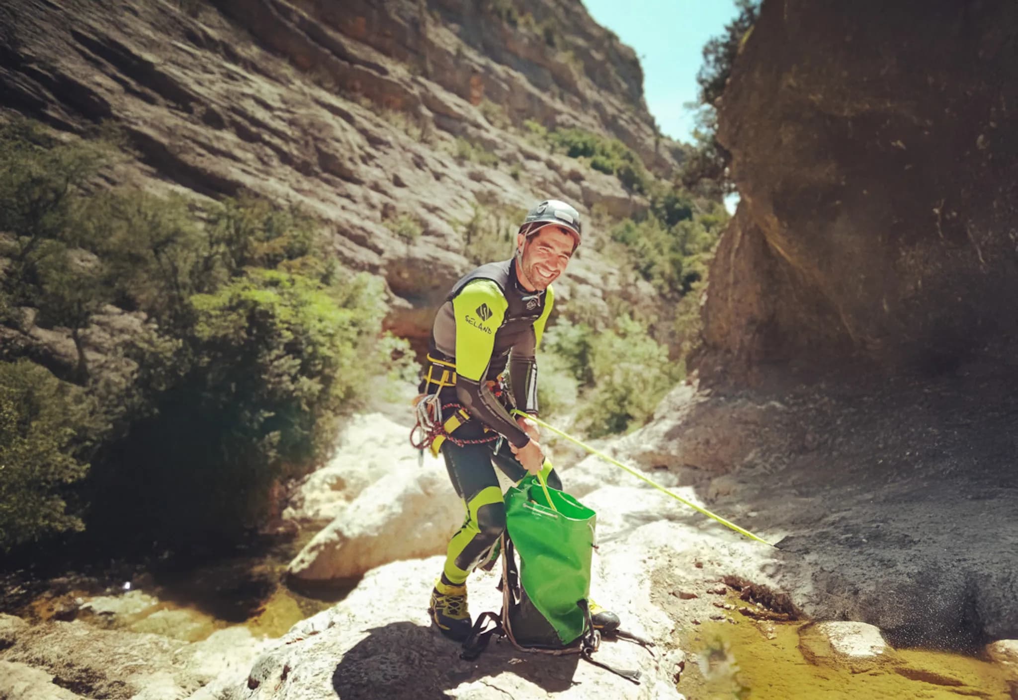 A smiling man stands on a rocky surface, wearing a black and green canyoning suit. He is holding a green bag and appears to be preparing his equipment, with a rope tied around his waist. The landscape in the background shows