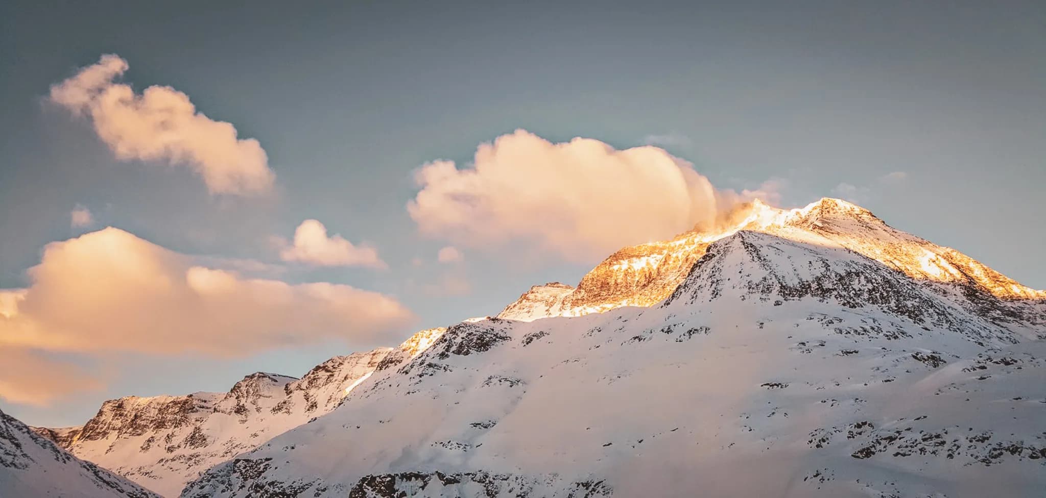 A snow-capped mountain peak illuminated by the golden light of sunset, with light clouds floating in the sky. The steep slopes and jutting ridges add to the majesty of the