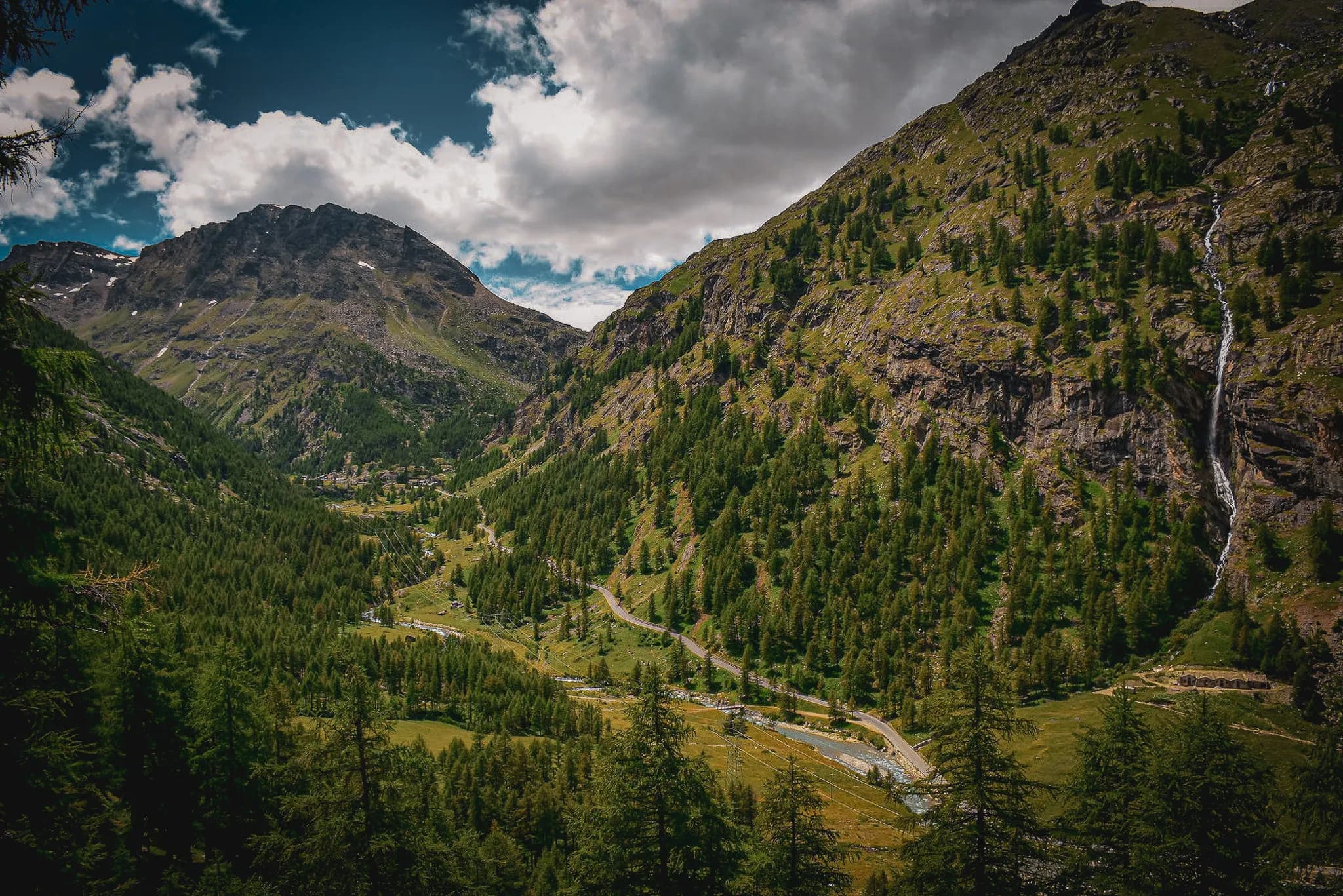 Een spectaculair berglandschap met steile, groene toppen. In het midden strekt zich een weelderige vallei uit, omzoomd door naaldbomen en doorkruist door kronkelende paden.