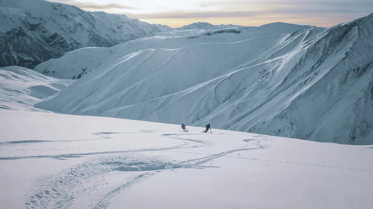 Deux skieurs s'élancent sur des pentes enneigées, illuminées par un doux coucher de soleil.