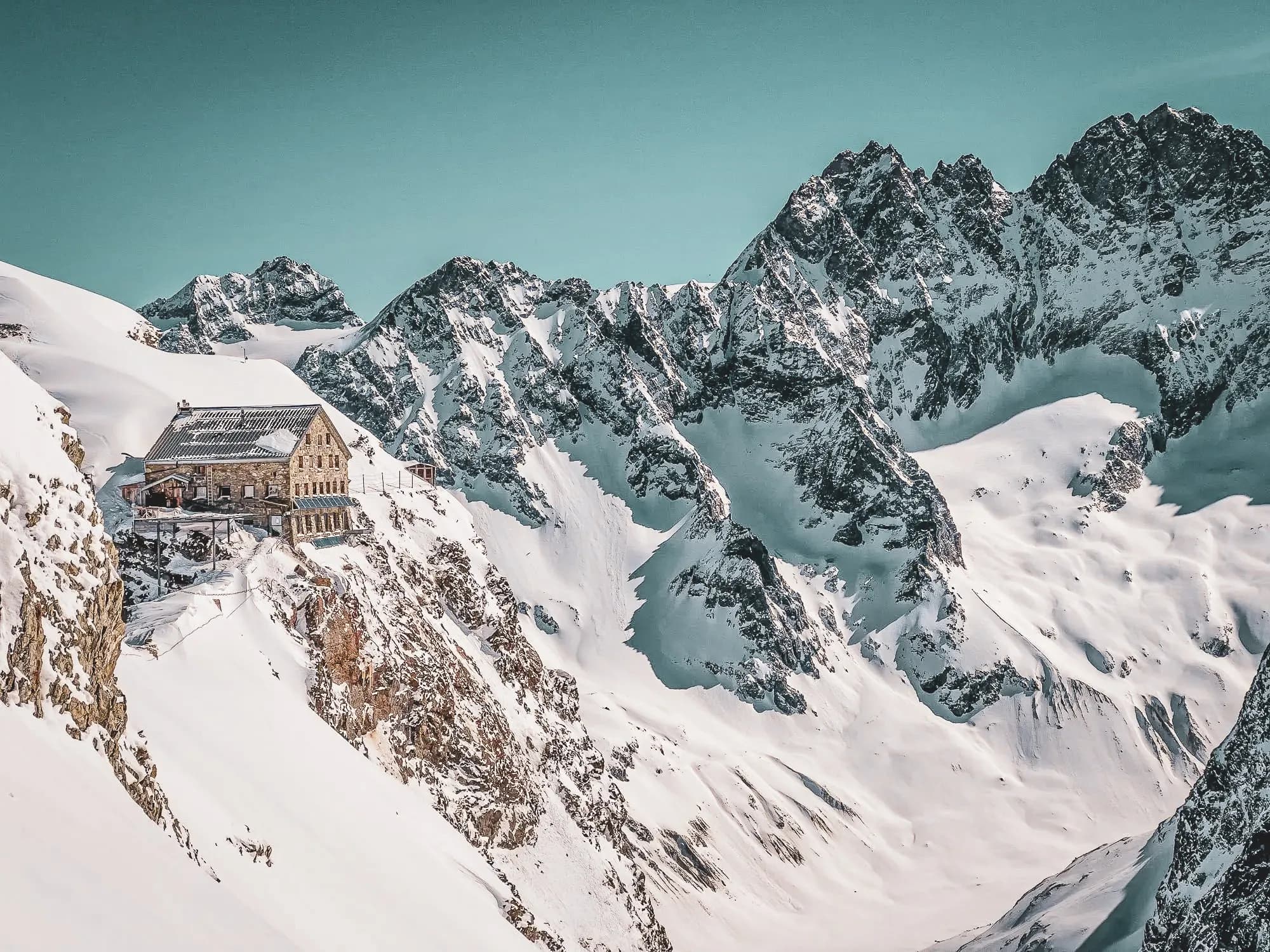 A stone mountain hut on a snow-covered peak, surrounded by mountain scenery. Stairs lead up to the refuge, which has a metal awning. A skier carrying a rucksack