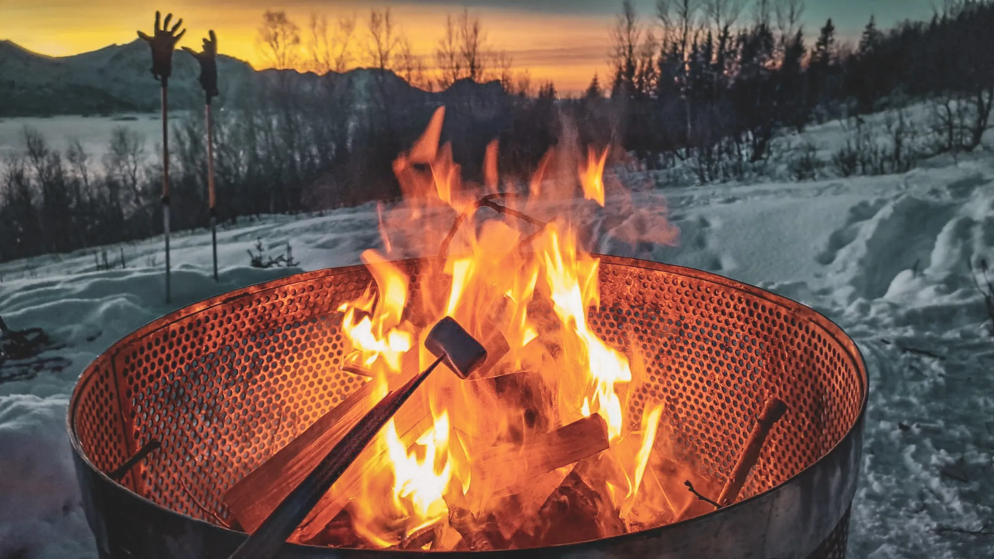 A warm fire crackles in a snowy landscape, with a sunset in the background.