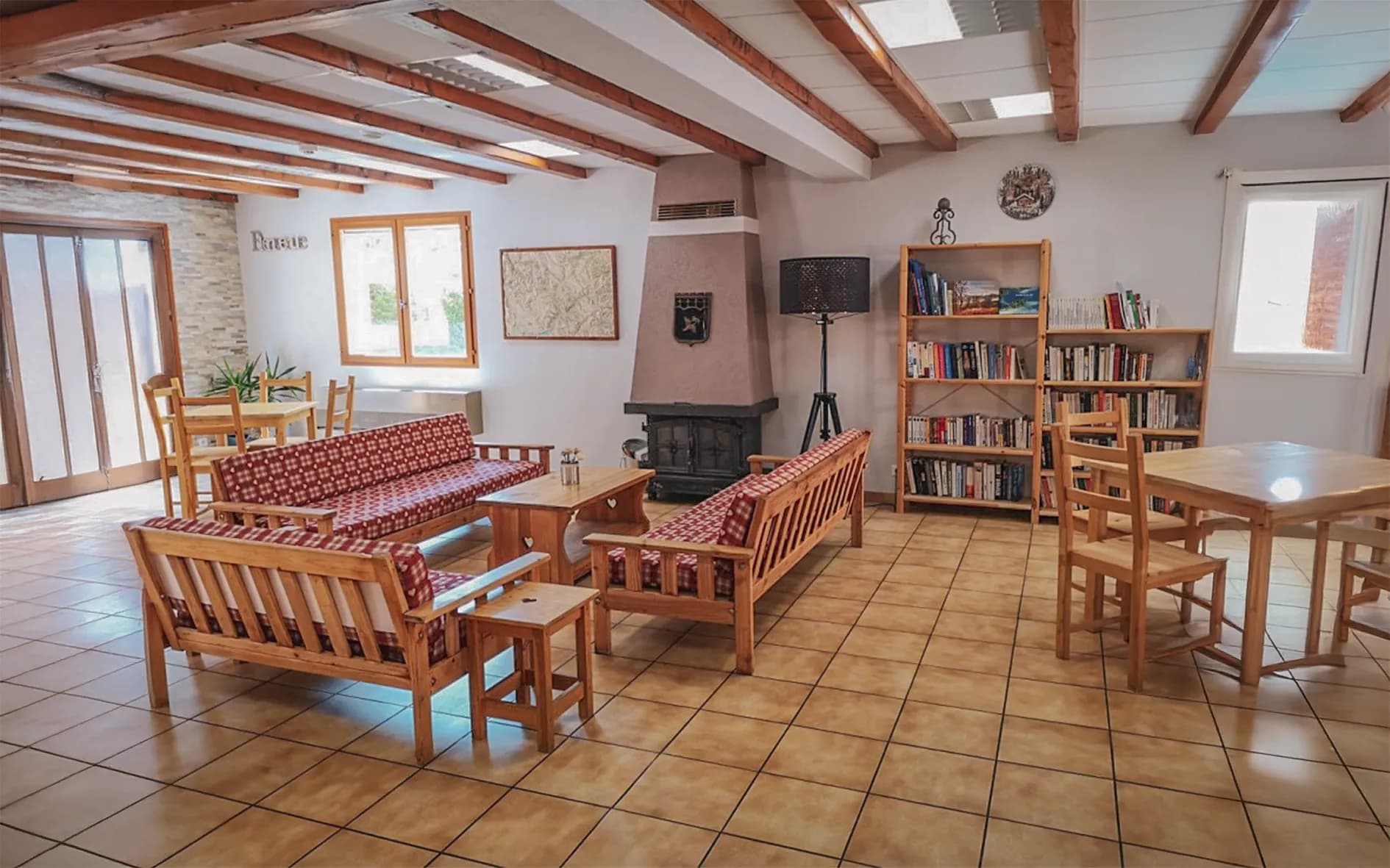 A cosy lounge with light-coloured walls and a beamed ceiling. Two wooden sofas with red cushions in a square pattern are arranged around a wooden table, accompanied by a small dining table.