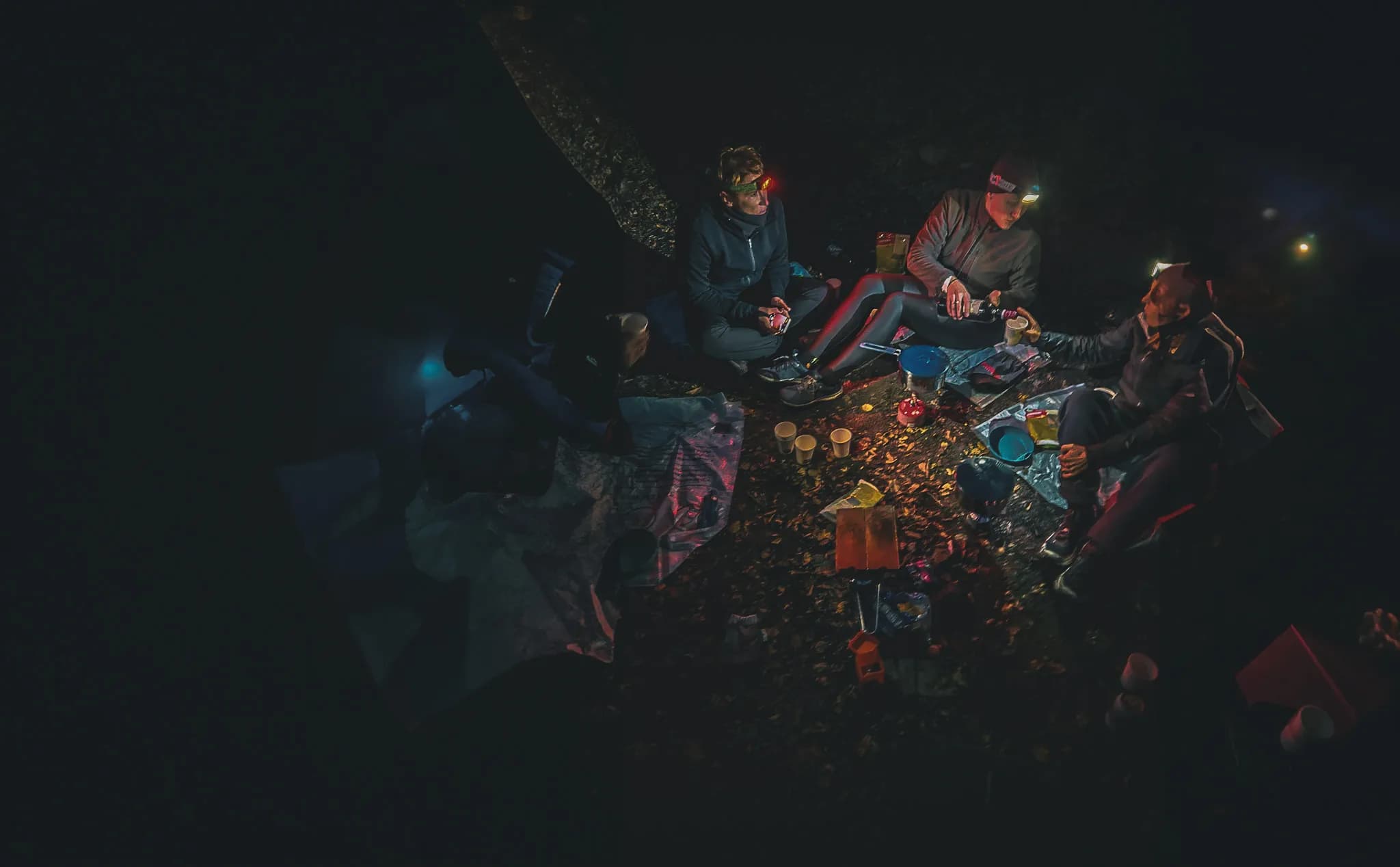 A group of four people are sitting on the ground, surrounded by leaves and dark night. Some of them are carrying lit headlamps, illuminating the space around them. On the ground, there are various utensils