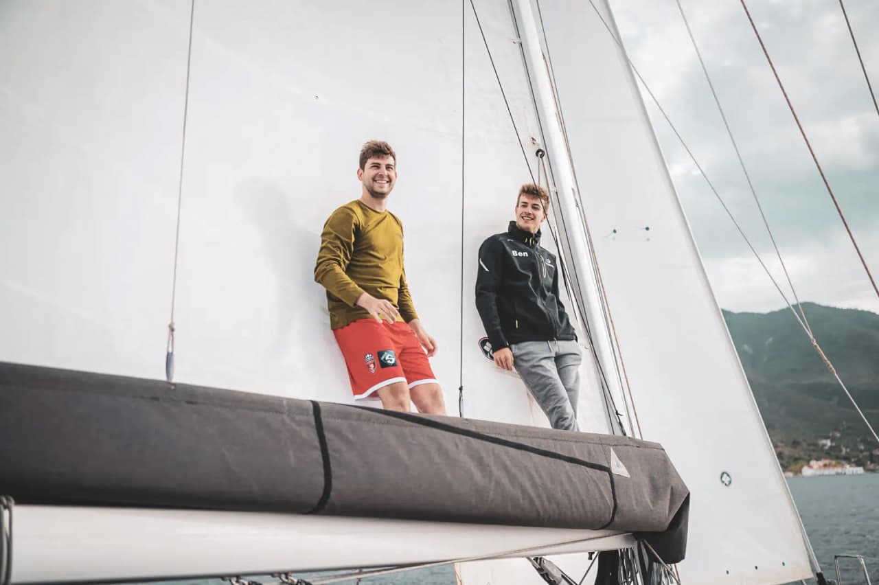 Deux jeunes hommes souriants profitent d'une balade en voilier, entourés par la mer et les montagnes.