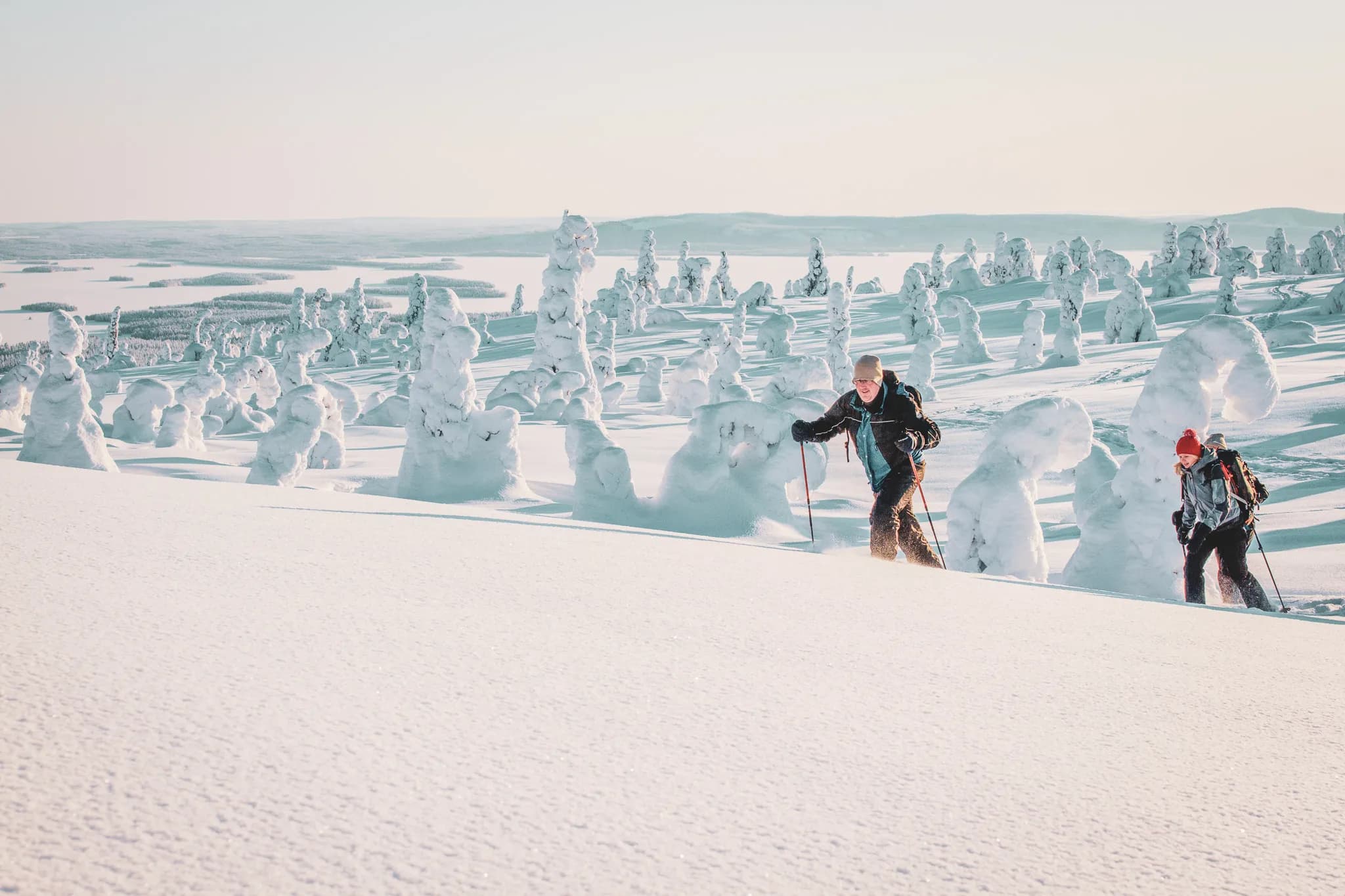 A snow-covered landscape with silhouettes of fir trees covered in snow, creating an enchanting atmosphere. Two hikers, wearing winter clothes, walk up a slope. One of them is using