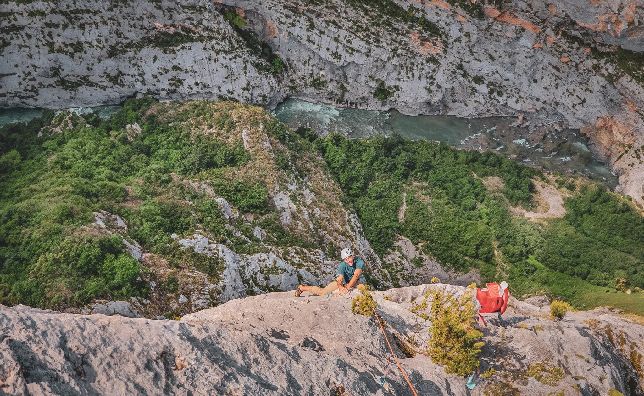 A climber is suspended on the edge of a rocky cliff, with a bird's eye view of a verdant valley. Below, a rushing river winds its way through the landscape. Rocks and
