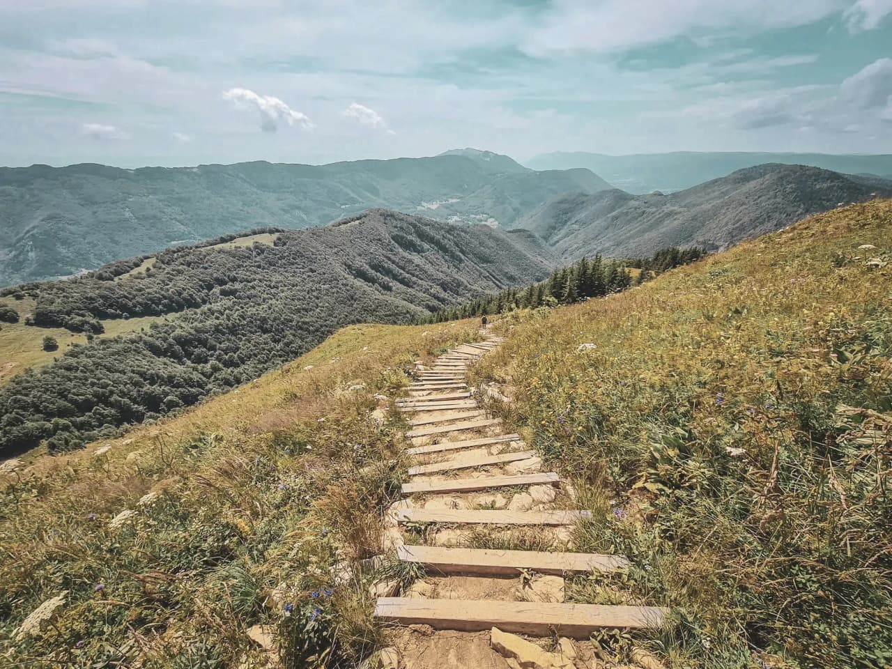 A wooden path winding through lush green mountains under a bright blue sky.