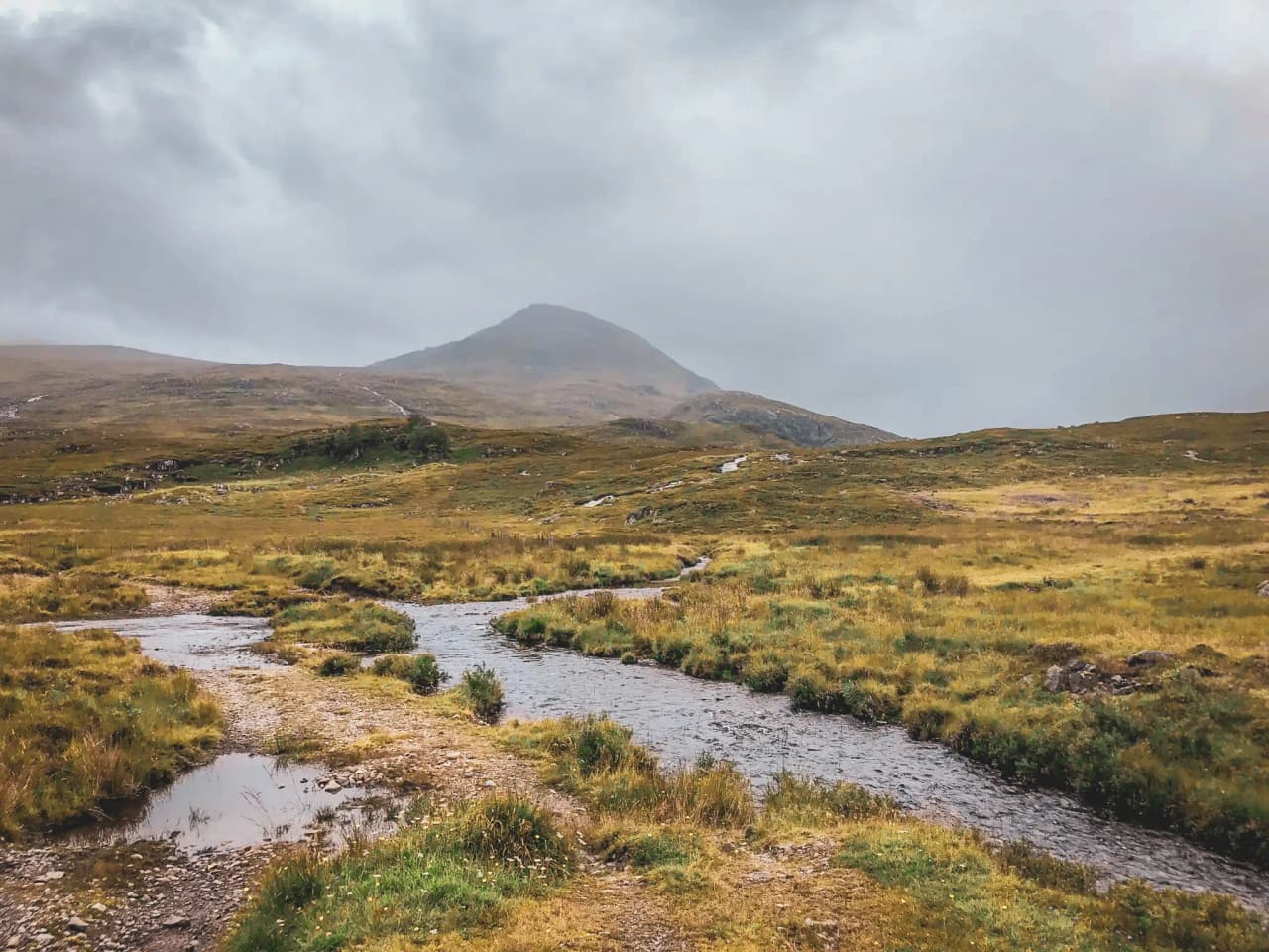 Een landschap van de Schotse Hooglanden, weelderig groene valleien en kronkelende beekjes onder een bewolkte hemel.