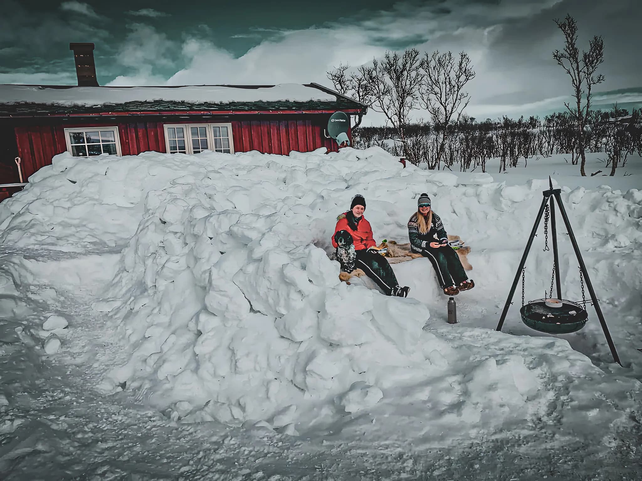 Two people enjoying a meal in the open air, surrounded by snow, near a red Mountain hut in Lapland.