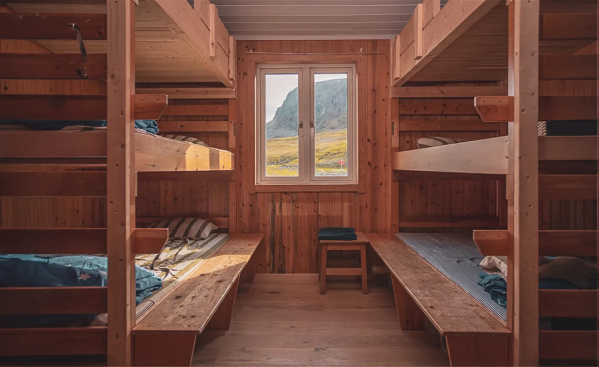 A wooden bedroom with bunk beds, offering a warm, rustic feel. The walls and furniture are made of wood, creating a natural space. A window in the centre overlooks a mountain landscape, illuminating the room.