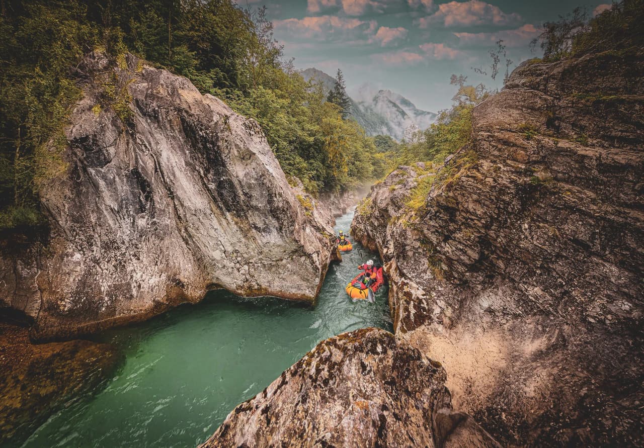 A turquoise stream winds its way through towering rock formations, surrounded by dense vegetation. Adventurers in orange kayaks glide across the water, navigating a narrow passage between two large rocks. Visit