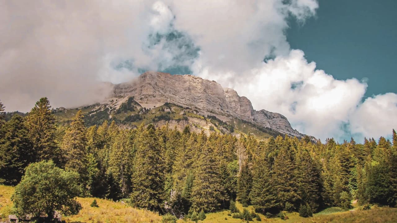 Majestueuse montagne éclairée par le soleil, entourée de forêts verdoyantes et de nuages.