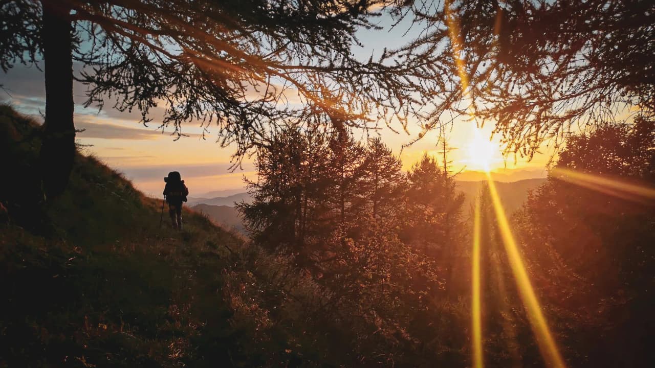 Une silhouette de randonneur sur un sentier, face à un coucher de soleil éclatant en montagne.