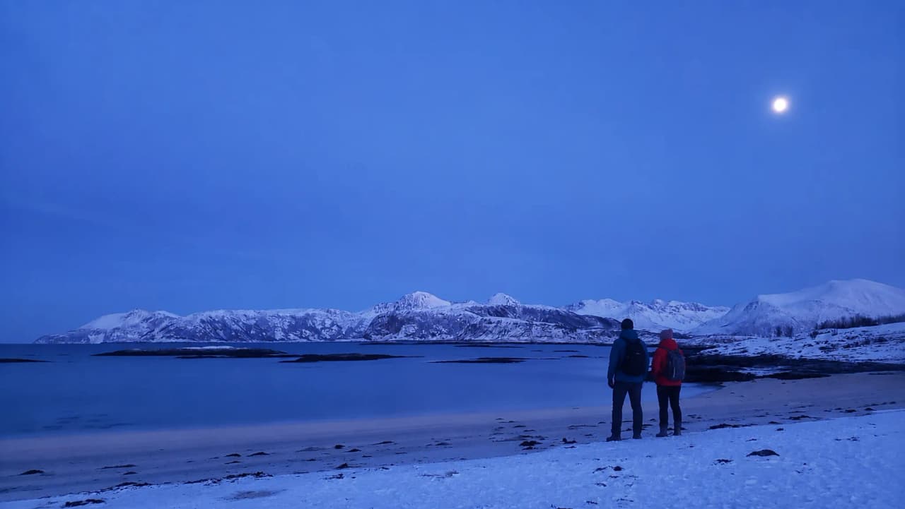 Twee wandelaars bewonderen bij schemering een besneeuwd landschap, met uitzicht op de zee en de bergen.