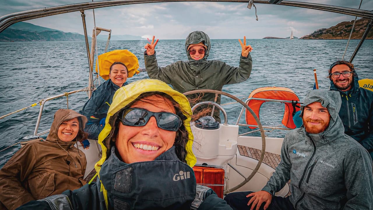 Een lachende groep op een zeilboot in de Middellandse Zee, onder een bewolkte hemel, met een avontuurlijke sfeer.