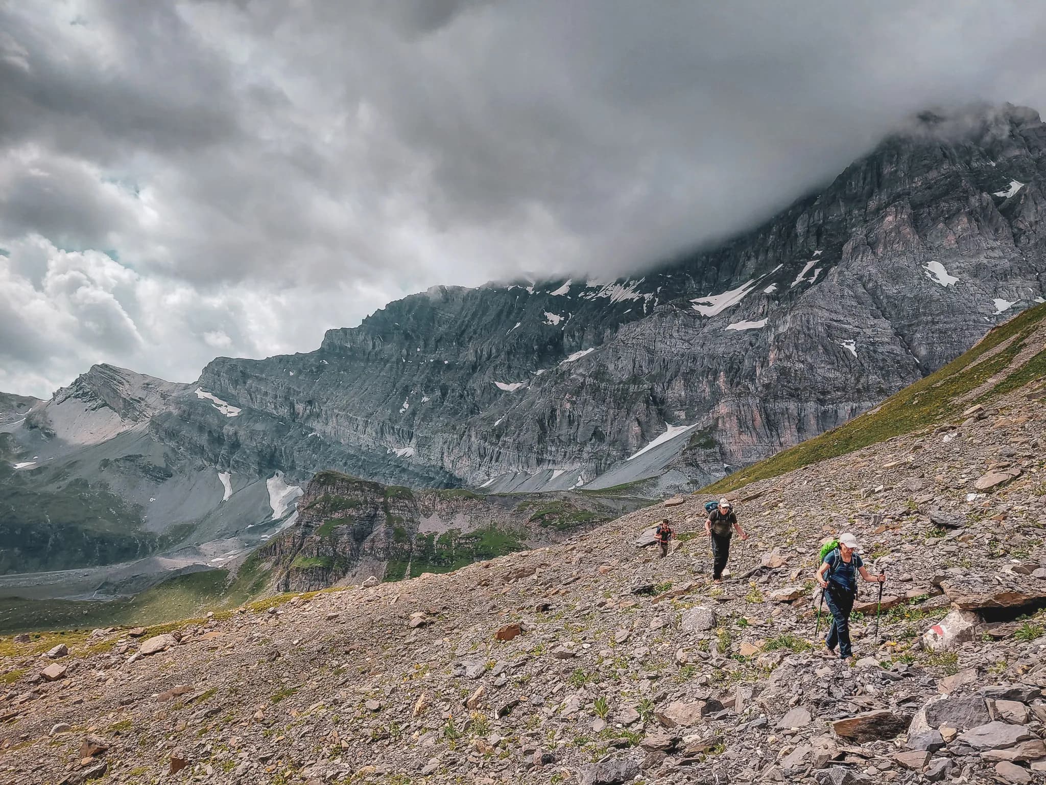 A spectacular mountain landscape, characterised by steep peaks and well-defined rocky ridges, with shades of grey and brown, interspersed with traces of snow. The sky is partly cloudy