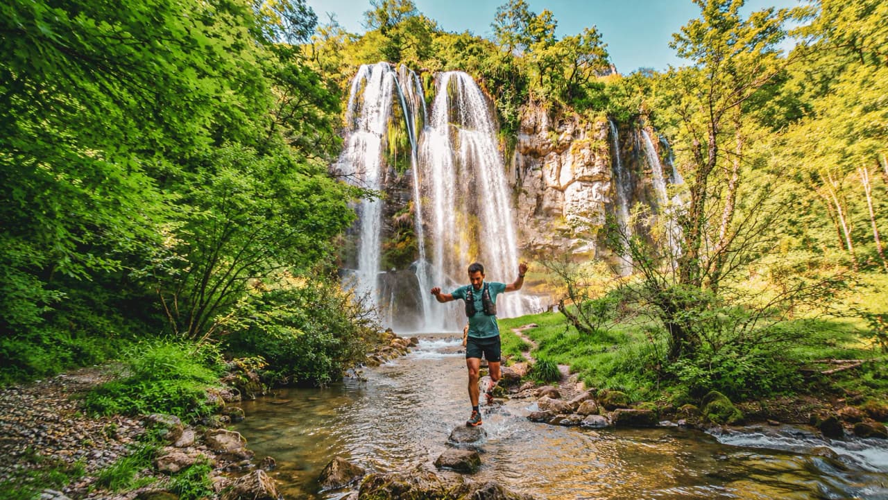 A runner crossing a stream in front of a majestic green waterfall, an invitation to adventure.