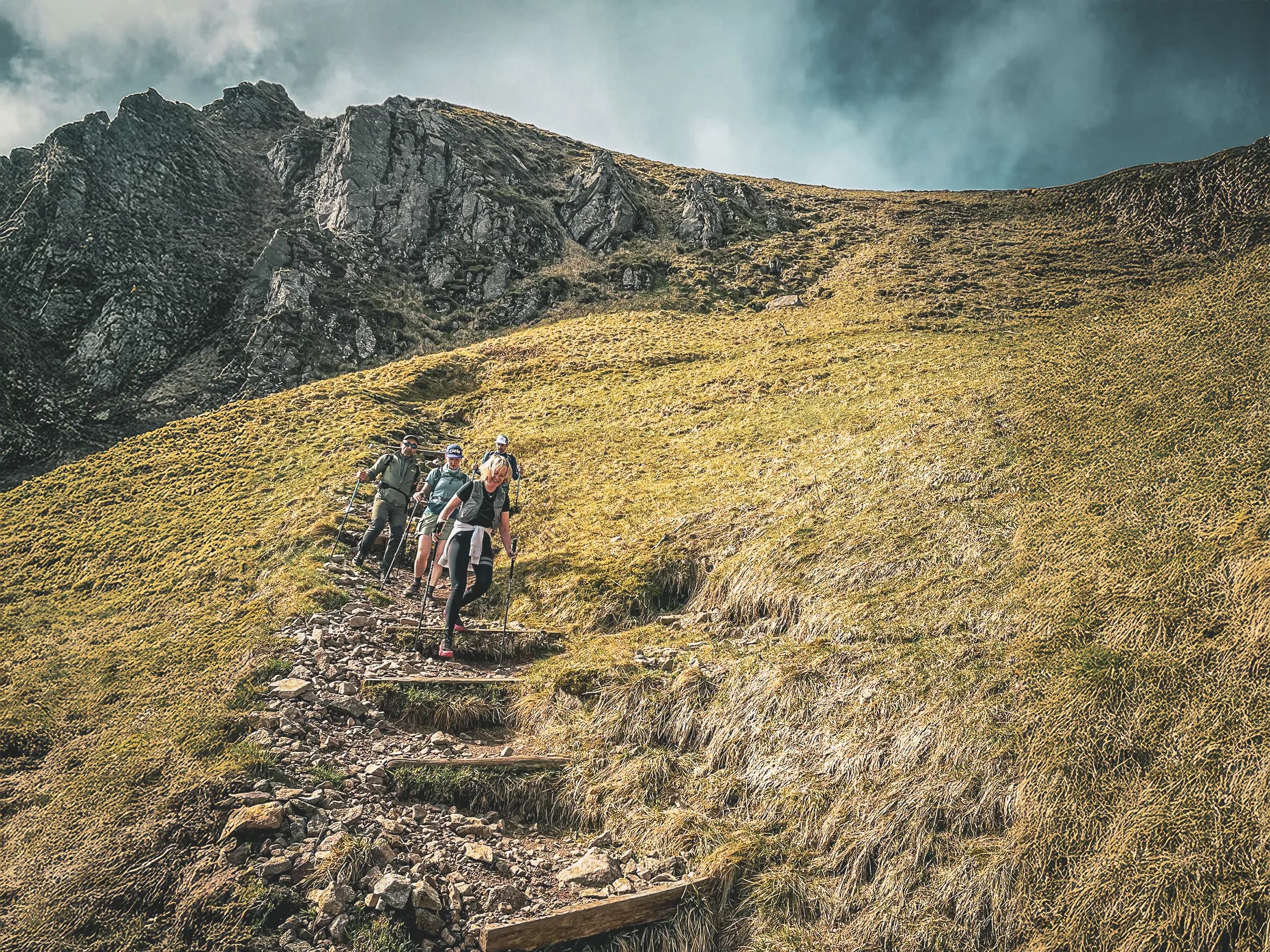 A group of runners trekking in the Vosges, climbing a green path under cloudy skies.