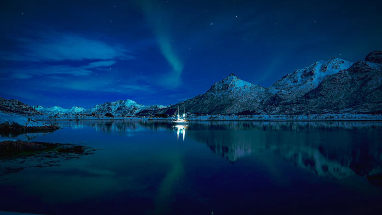 An illuminated sailing boat in a fjord at night, with snow-capped mountains and a starry sky in the background.