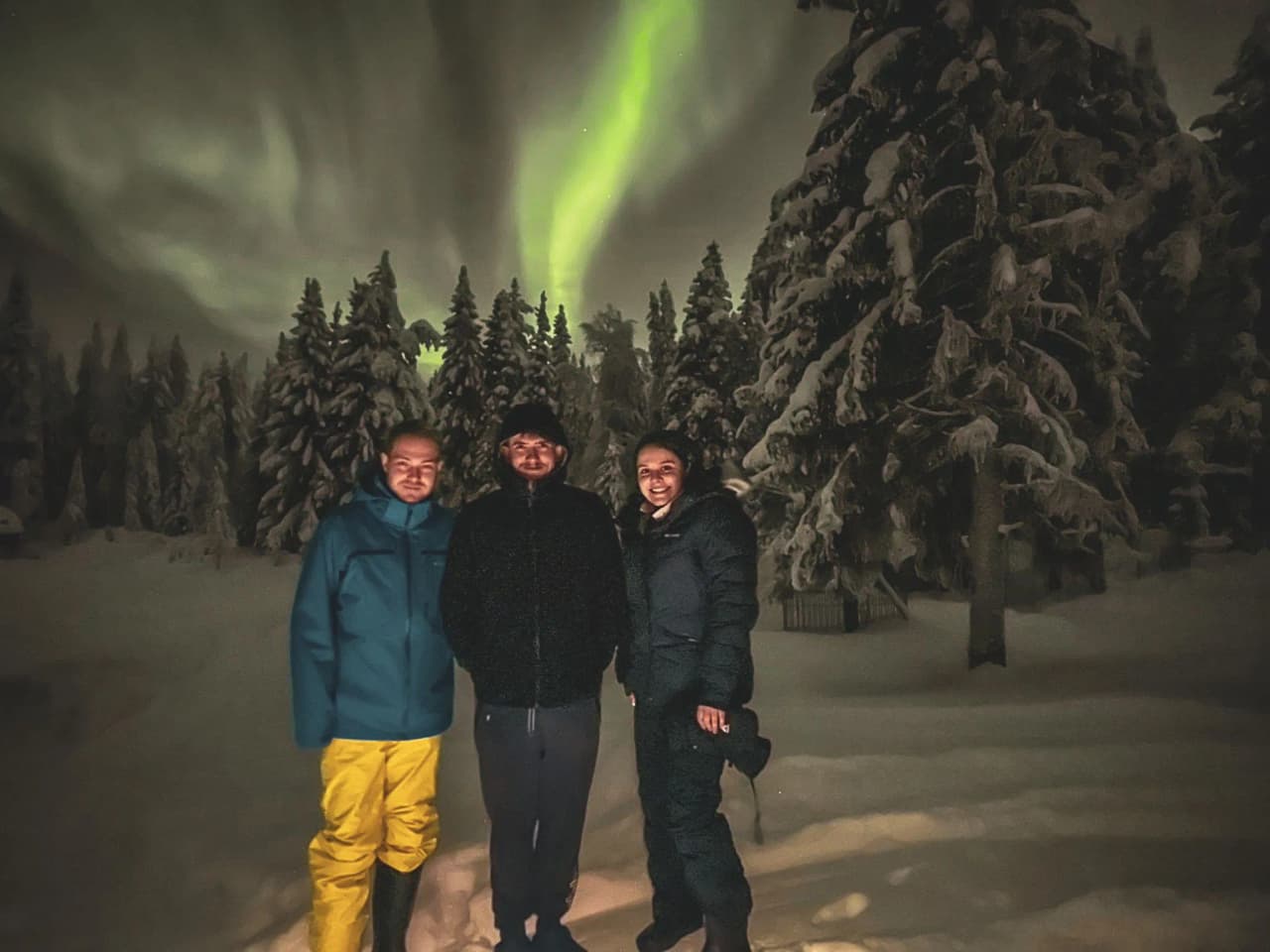 Drie lachende mensen onder het noorderlicht, omringd door besneeuwde landschappen in Lapland.