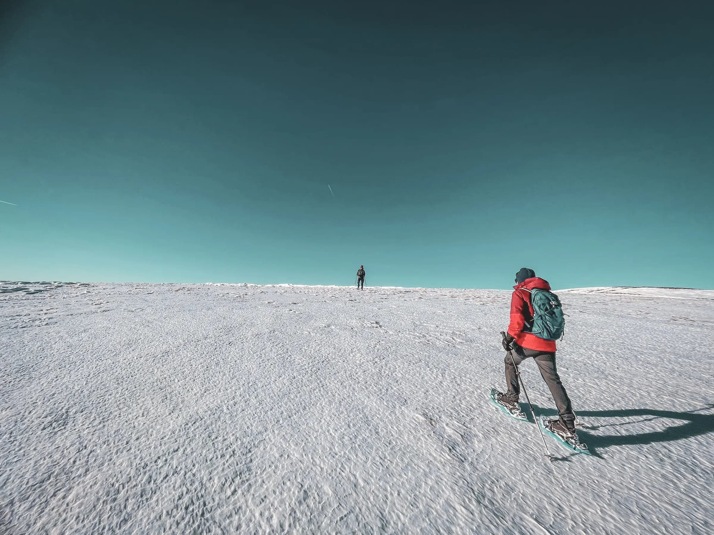 Een besneeuwd landschap strekt zich uit zover het oog reikt, met een intens blauwe lucht op de achtergrond. Twee mensen lopen over de sneeuw, de een bergopwaarts en de ander iets verderop, op wat glooiend terrein lijkt.