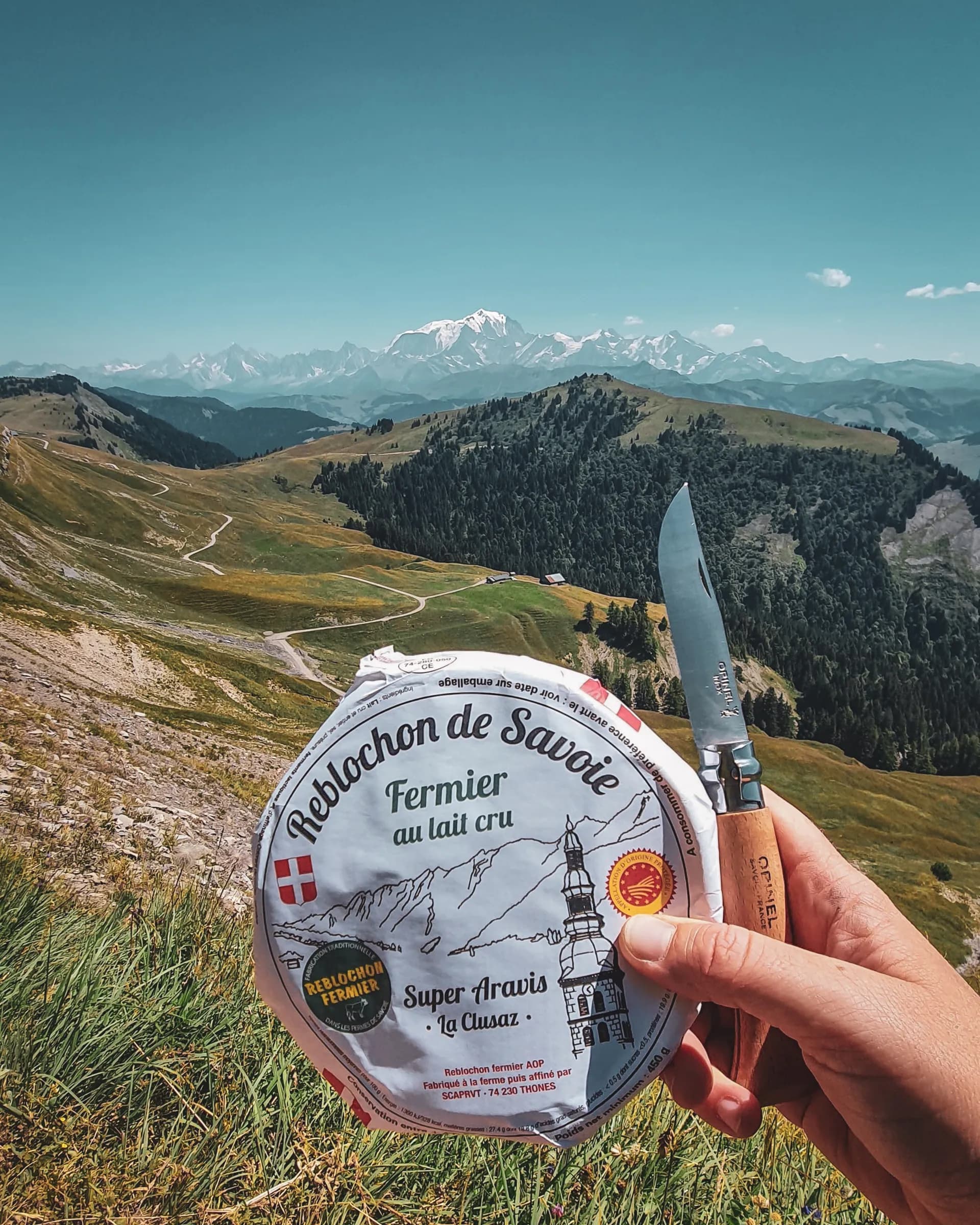 A hiker holds a Savoie Reblochon cheese with the Alpine peaks in the background, inviting you to escape.