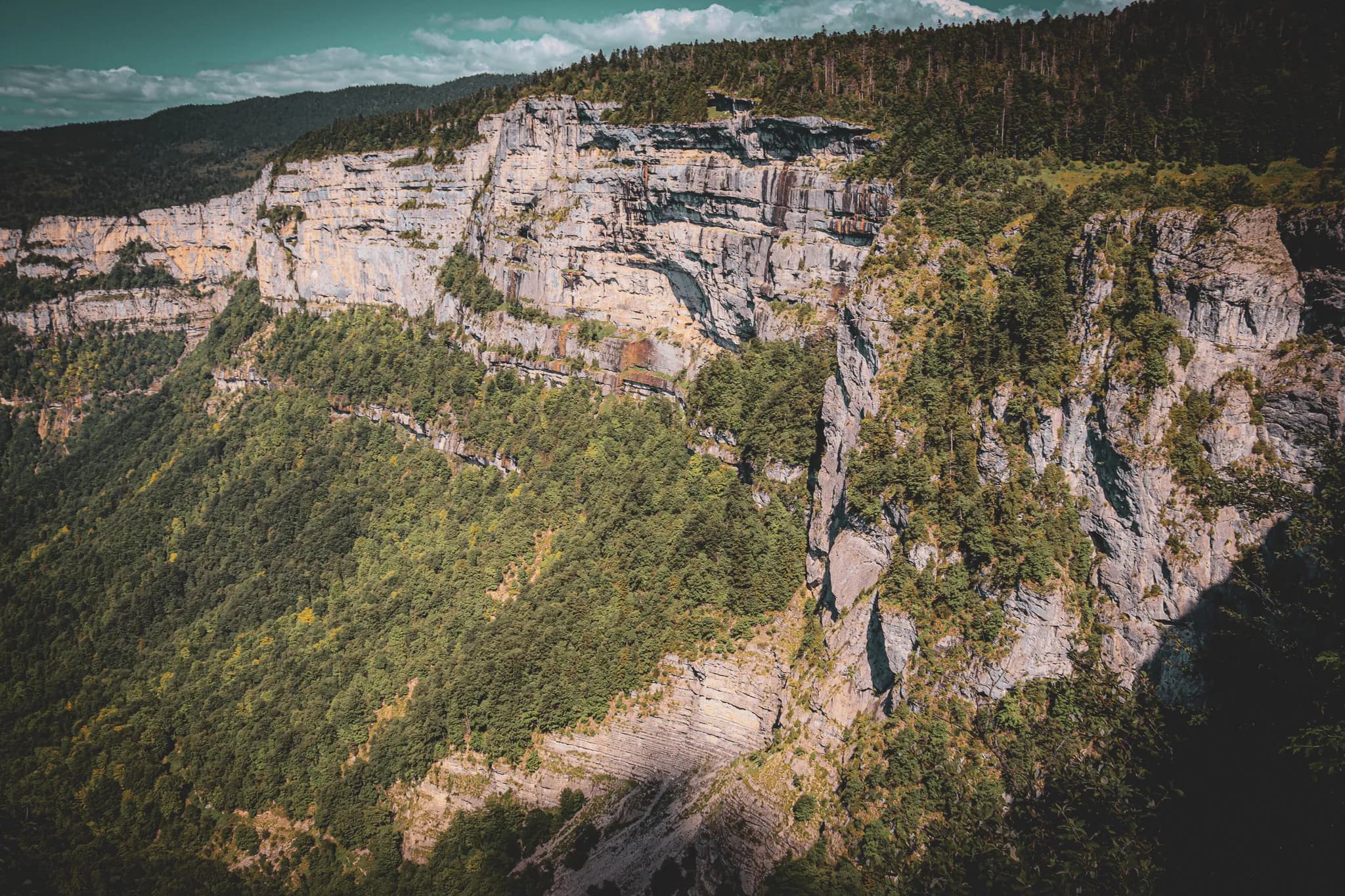 A mountainous landscape with steep cliffs surrounded by lush vegetation. The rocks are layered in shades of grey, while lush green trees line the base and slopes.