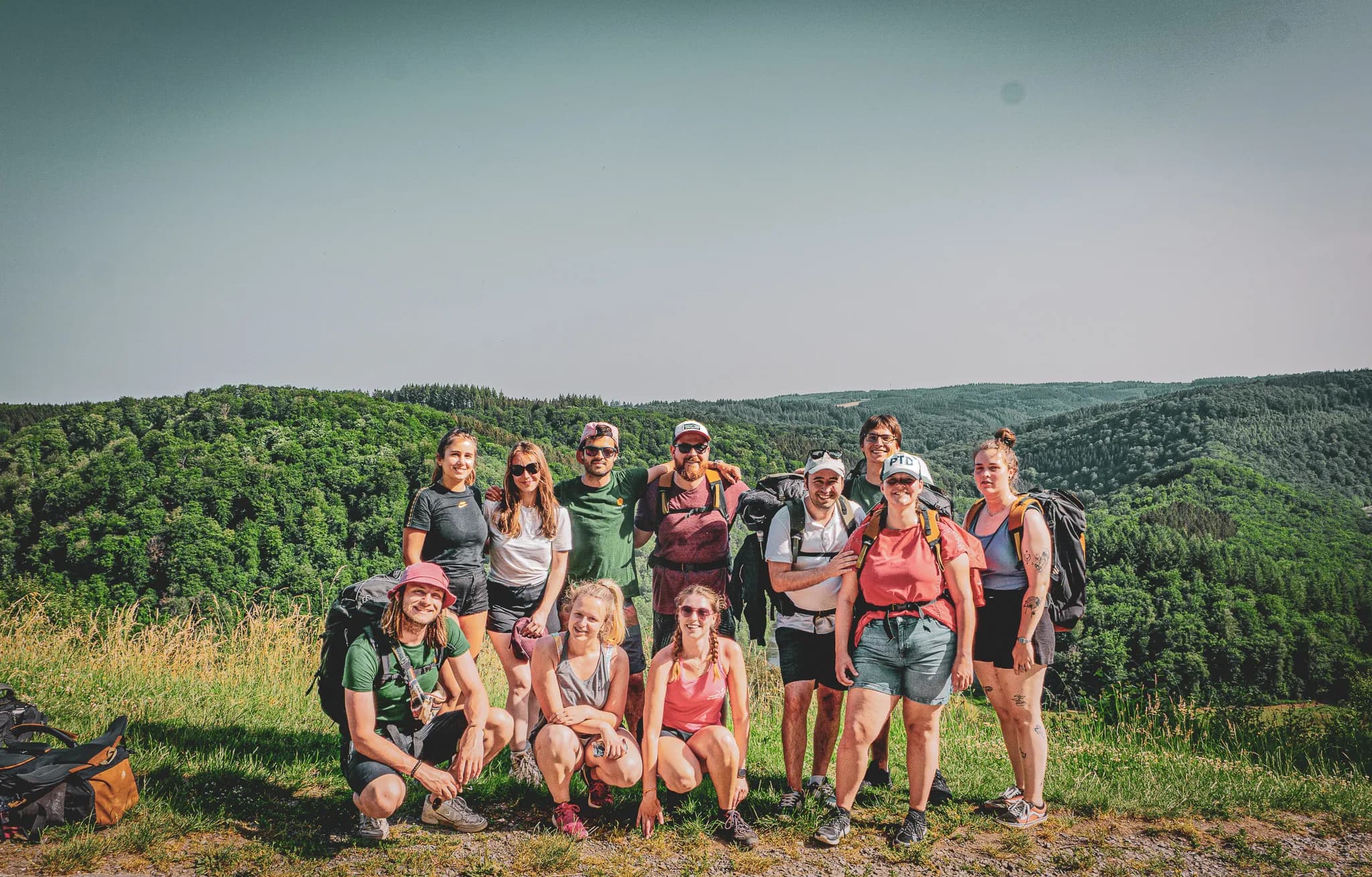 Een groep van twaalf mensen staat buiten en poseert tegen een achtergrond van groene heuvels onder een strakblauwe hemel. De meesten van de groep dragen t-shirts, shorts en petten die geschikt zijn voor de activiteiten waaraan ze gaan deelnemen.