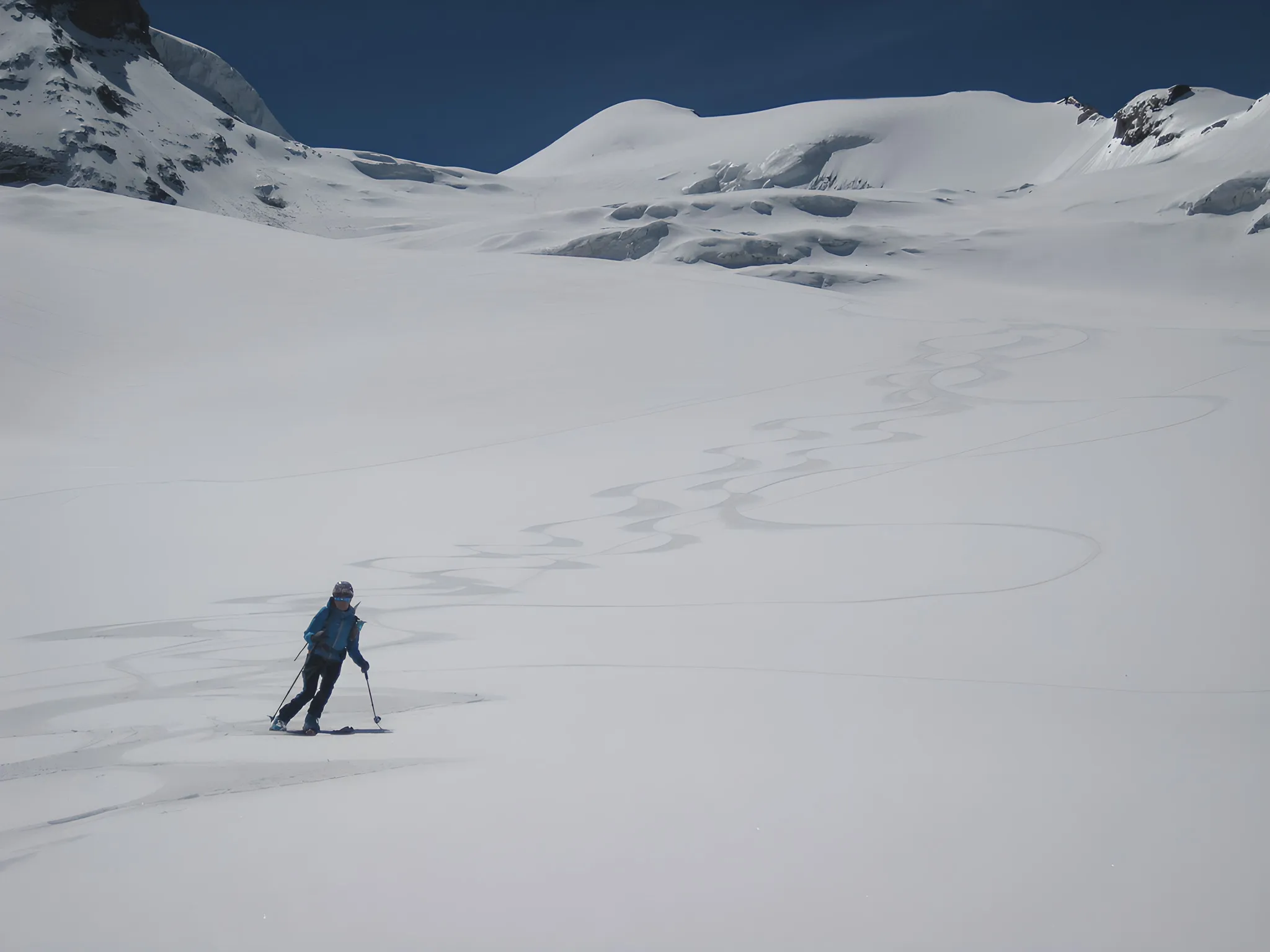 Un skieur solitaire trace des courbes élégantes sur un vaste glacier immaculé sous un ciel bleu.