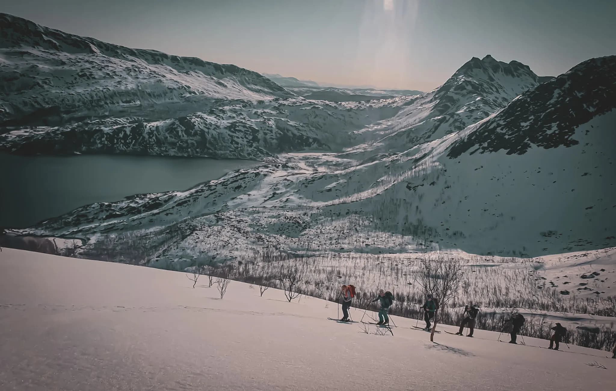 Een indrukwekkend berglandschap, met besneeuwde toppen erboven en een dal dat naar beneden glooit. Links weerspiegelt een rustig meer de omringende bergen. Op de voorgrond beklimt een groep wandelaars