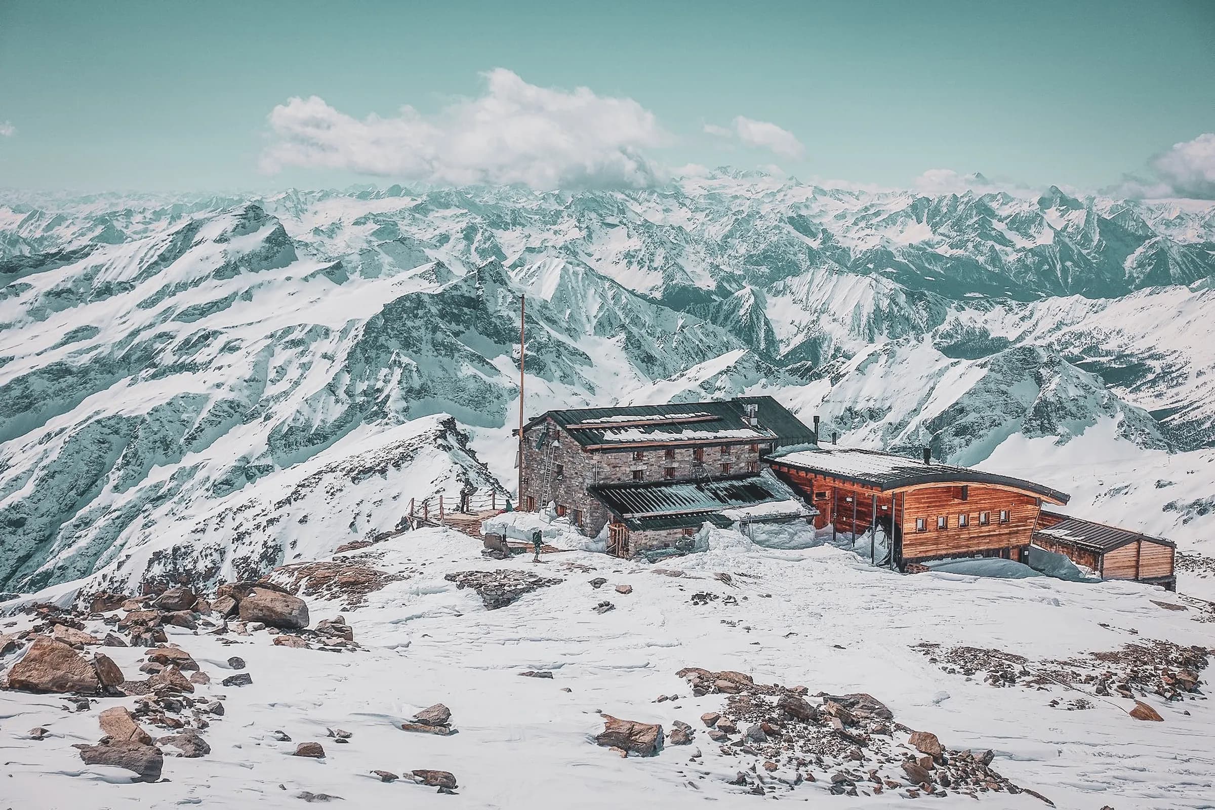 A wooden mountain chalet stands at the summit of a spectacular panorama. The refuge is surrounded by vast expanses of snow and majestic mountain ranges covered in glaciers. The sky is