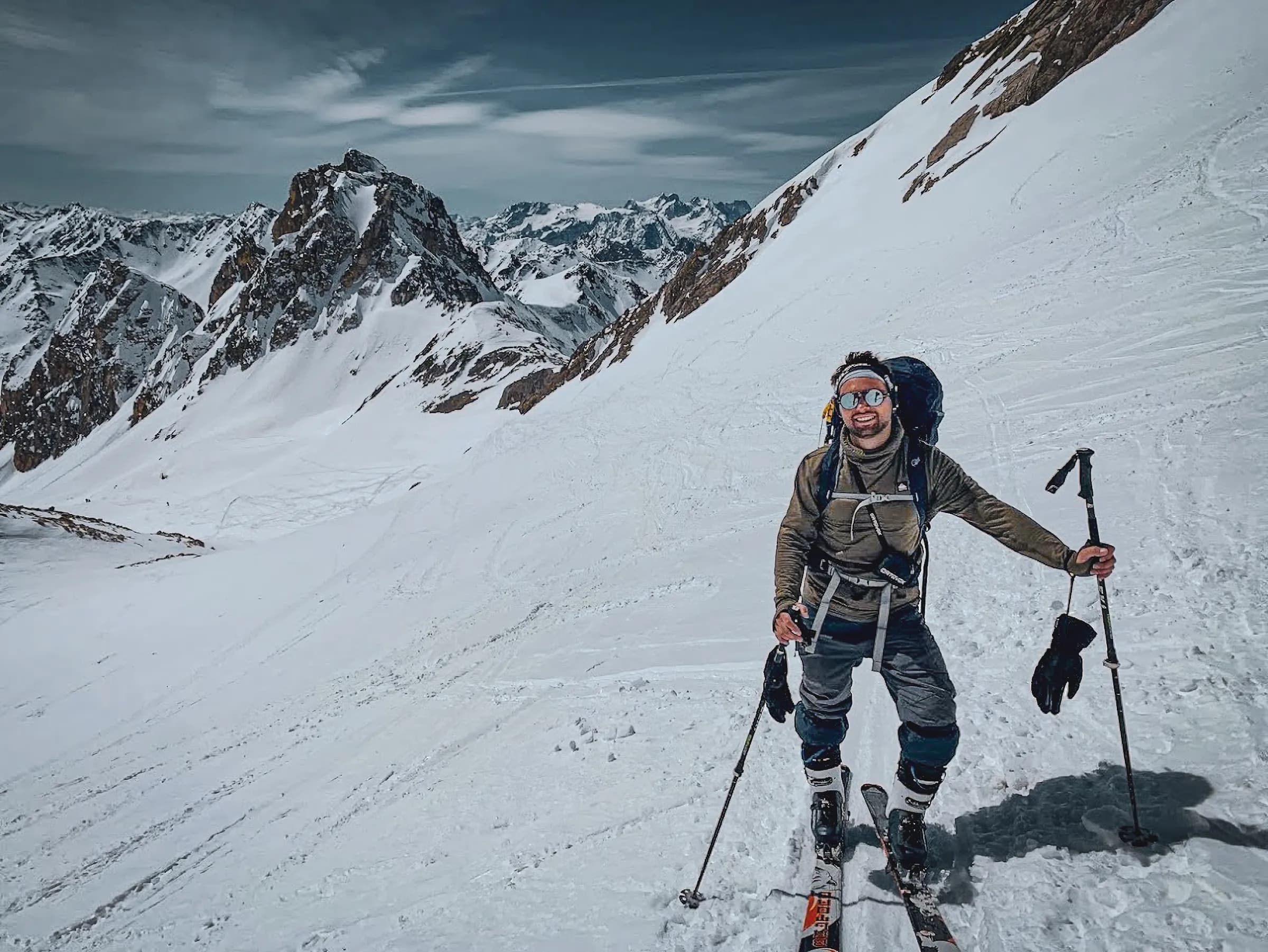 Un aventurier en montagne, souriant et équipé pour le ski de randonnée, se tient sur un terrain enneigé. Il porte des vêtements adaptés aux conditions froides, un sac à dos, et tient des bâtons de ski. En arrière