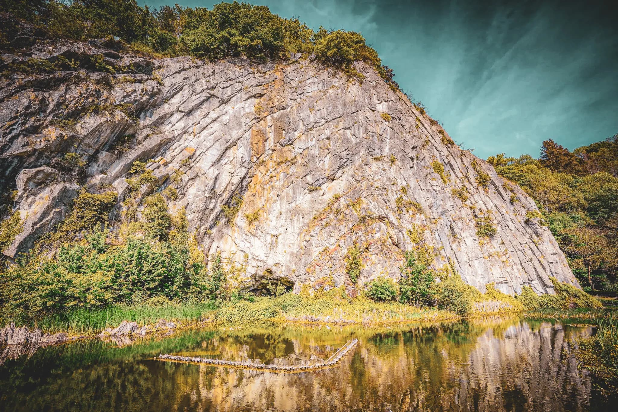 A captivating natural landscape with a rocky cliff on the left, covered in vegetation. The vertical wall is streaked and shades of grey, reflected in the calm water at its base. On the right,